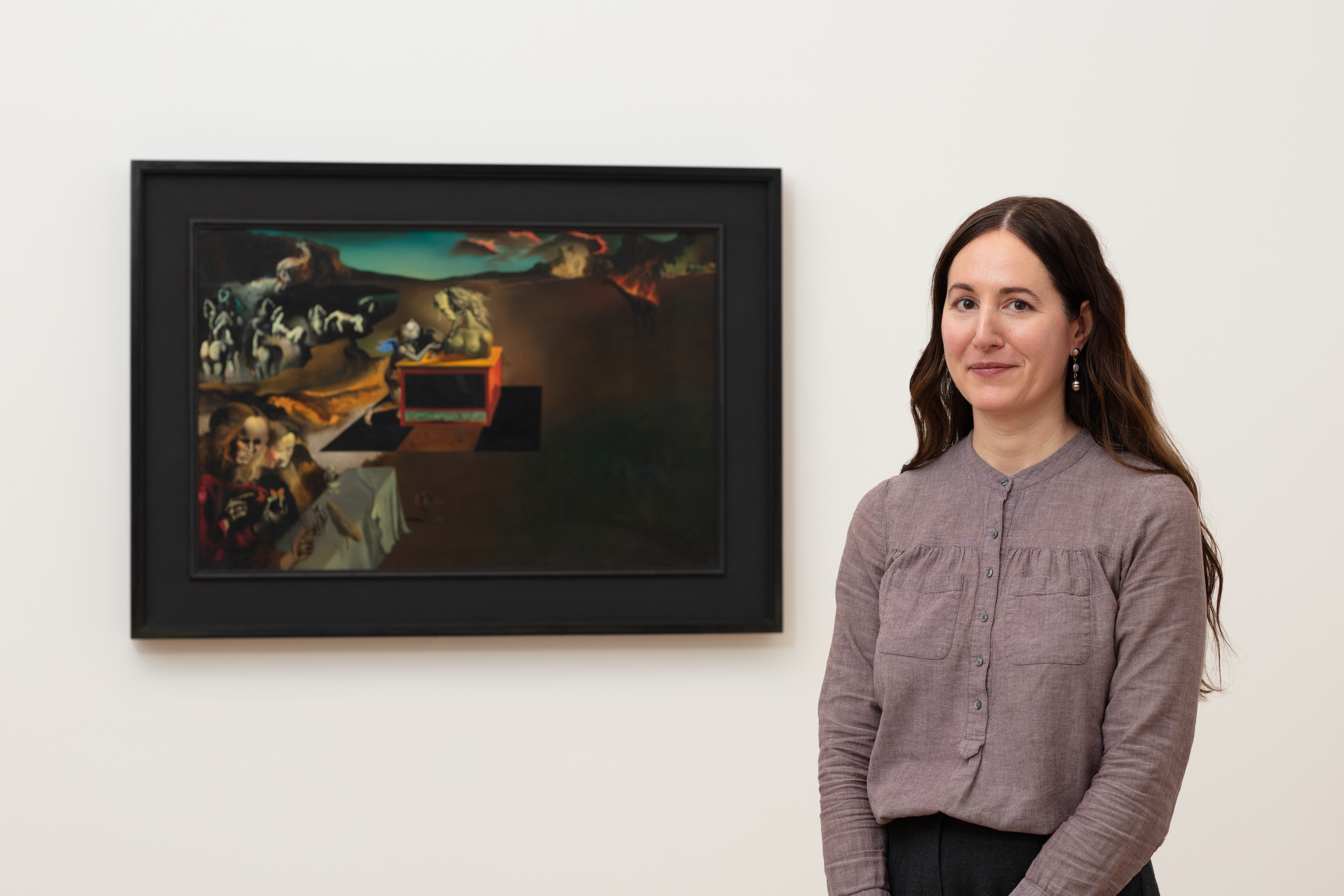 A light-skinned woman with light-brown hair in a gray linen shirt stands to the right of Salvador Dalí's painting "Inventions of the Monsters."
