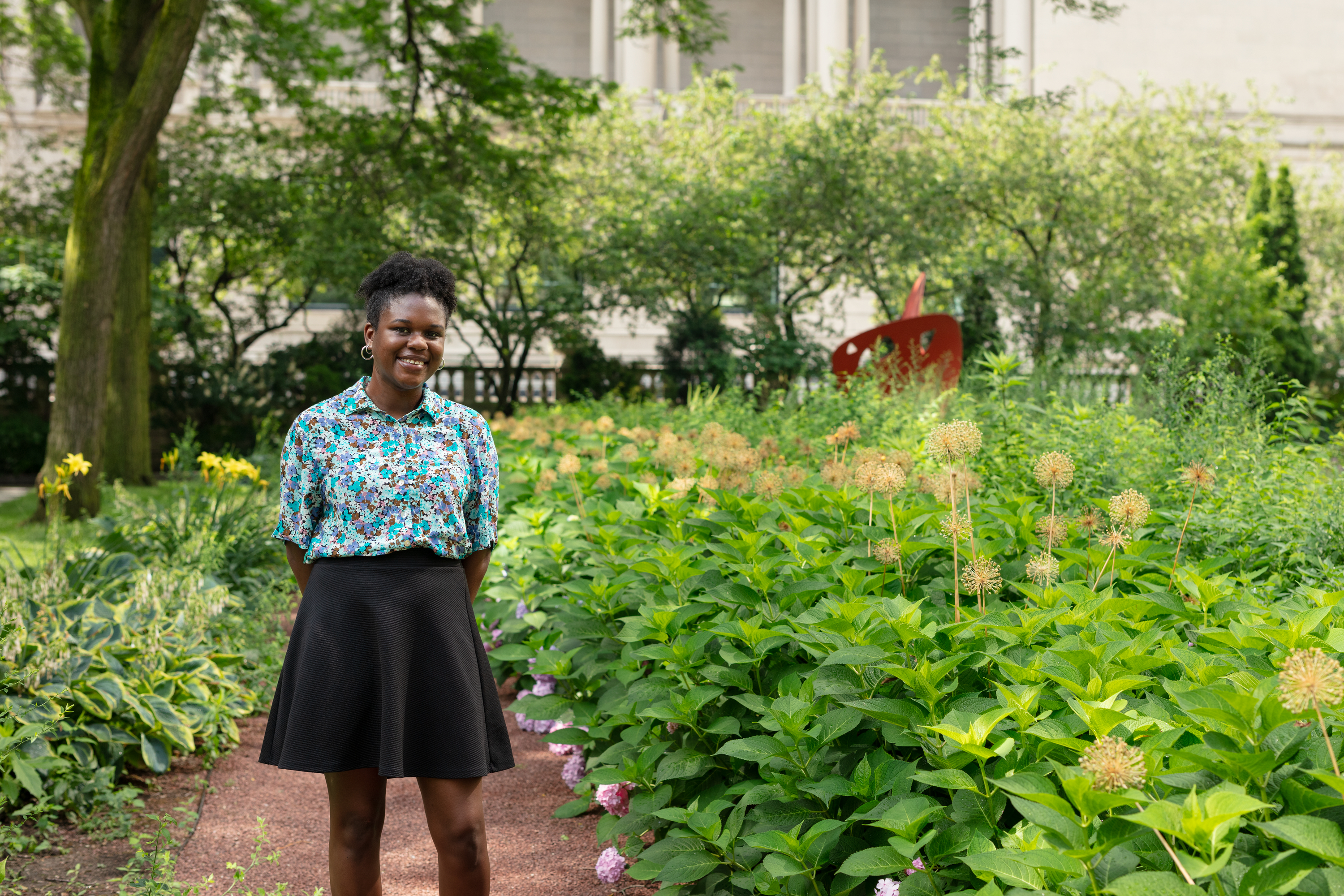 Photo of Loren Wright, a dark-skinned young woman in a bright floral top, standing in a lush, bright-green garden with a red sculpture in the background.