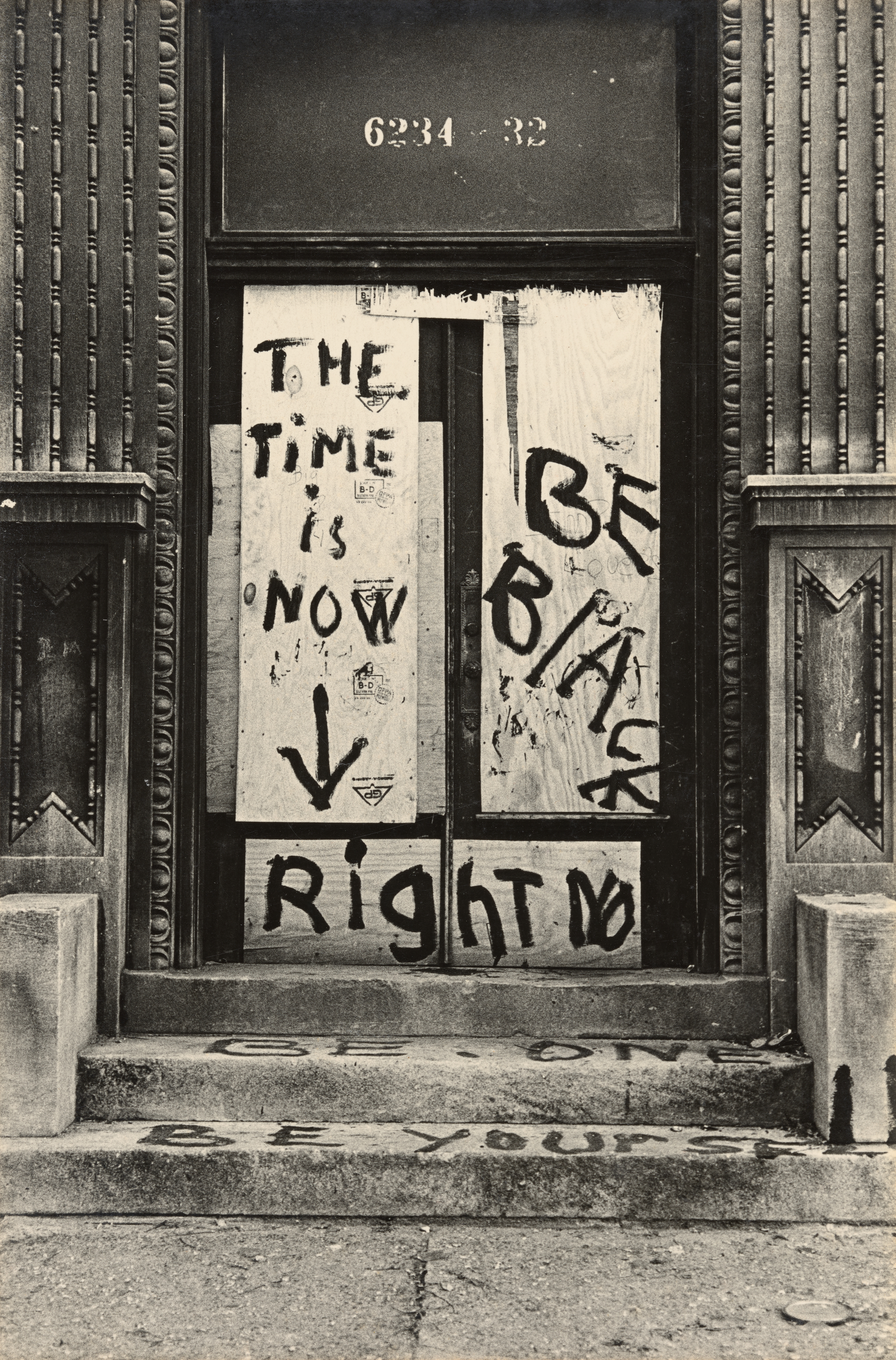 Black-and-white photograph of a boarded-up building entrance with stenciled address numbers “6234–32” above the doorway. The door is covered in plywood, painted with large, hand-lettered messages in dark paint. The text reads: “THE TIME IS NOW ↓,” “BE BLACK,” and “RIGHT NOW.” On the stone steps below, additional messages are painted: “BE ONE” and “BE YOURSELF.” The surrounding architectural details include ornate molding and decorative panels on either side of the doorway.