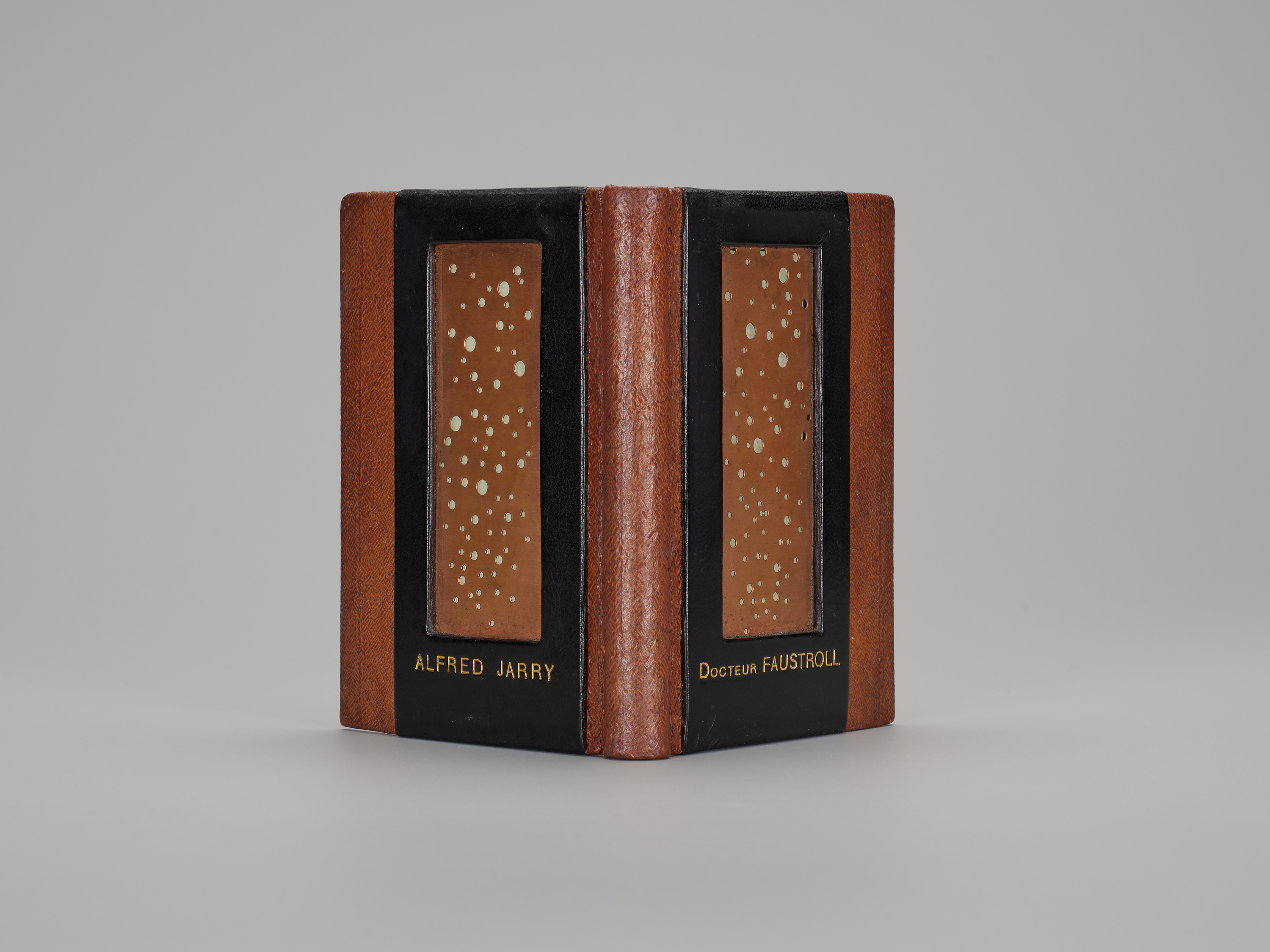 A hand-bound book, its spine facing the viewer and cover open. The book is bound in rich brown leather with a vertical, rectangular black frame inset in both covers. Within the black frame is a brown rectangle with many small circles cut out. The back cover reads "ALFRED JARRY," the front, "DOCTEUR FAUSTROLL."