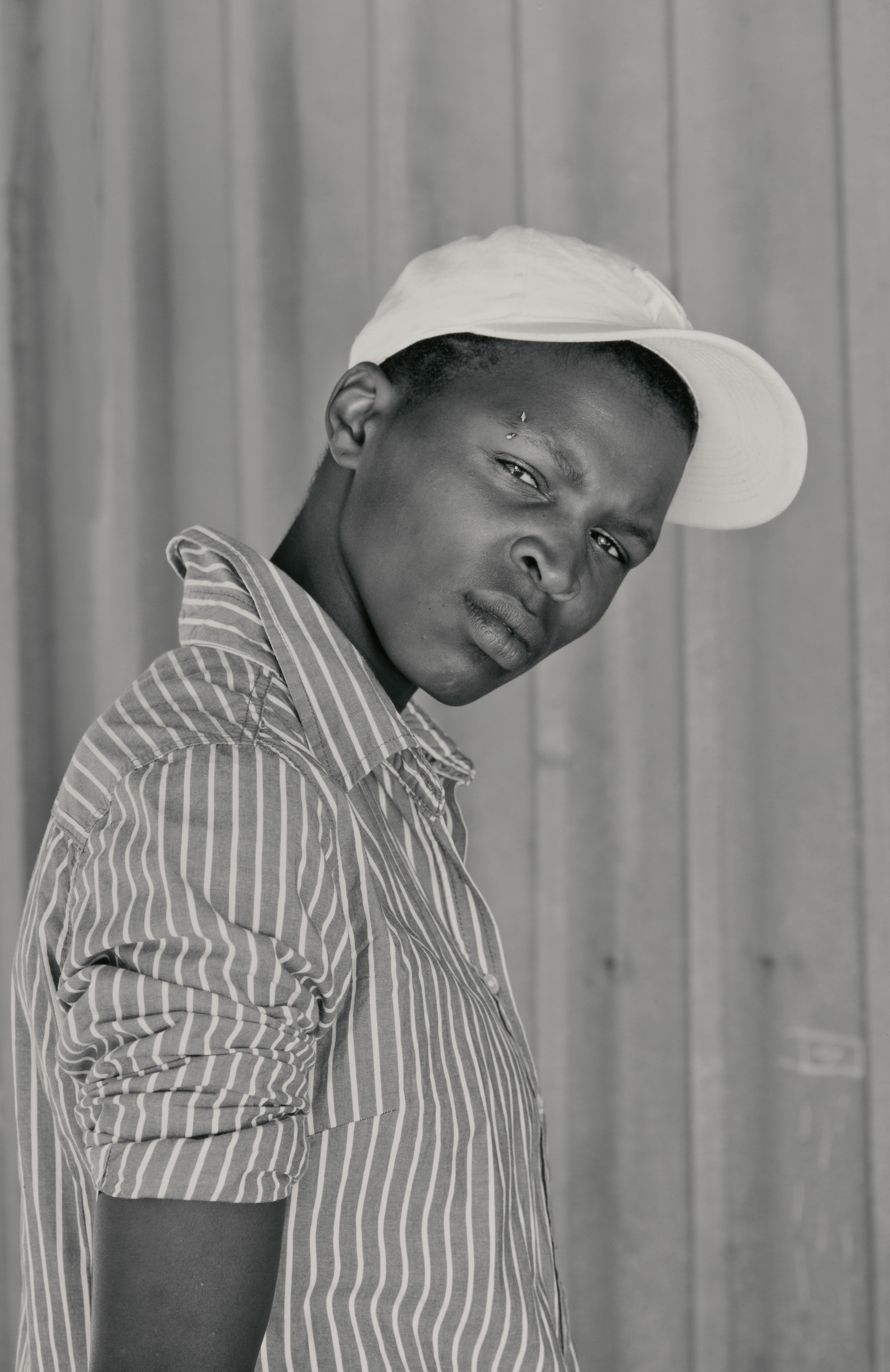 A black-and-white photograph of a dark-skinned person in profile with short hair wearing a light-colored cap and a striped collared shirt, their head tilted down and eyes looking straight into the camera with an almost defiant look.