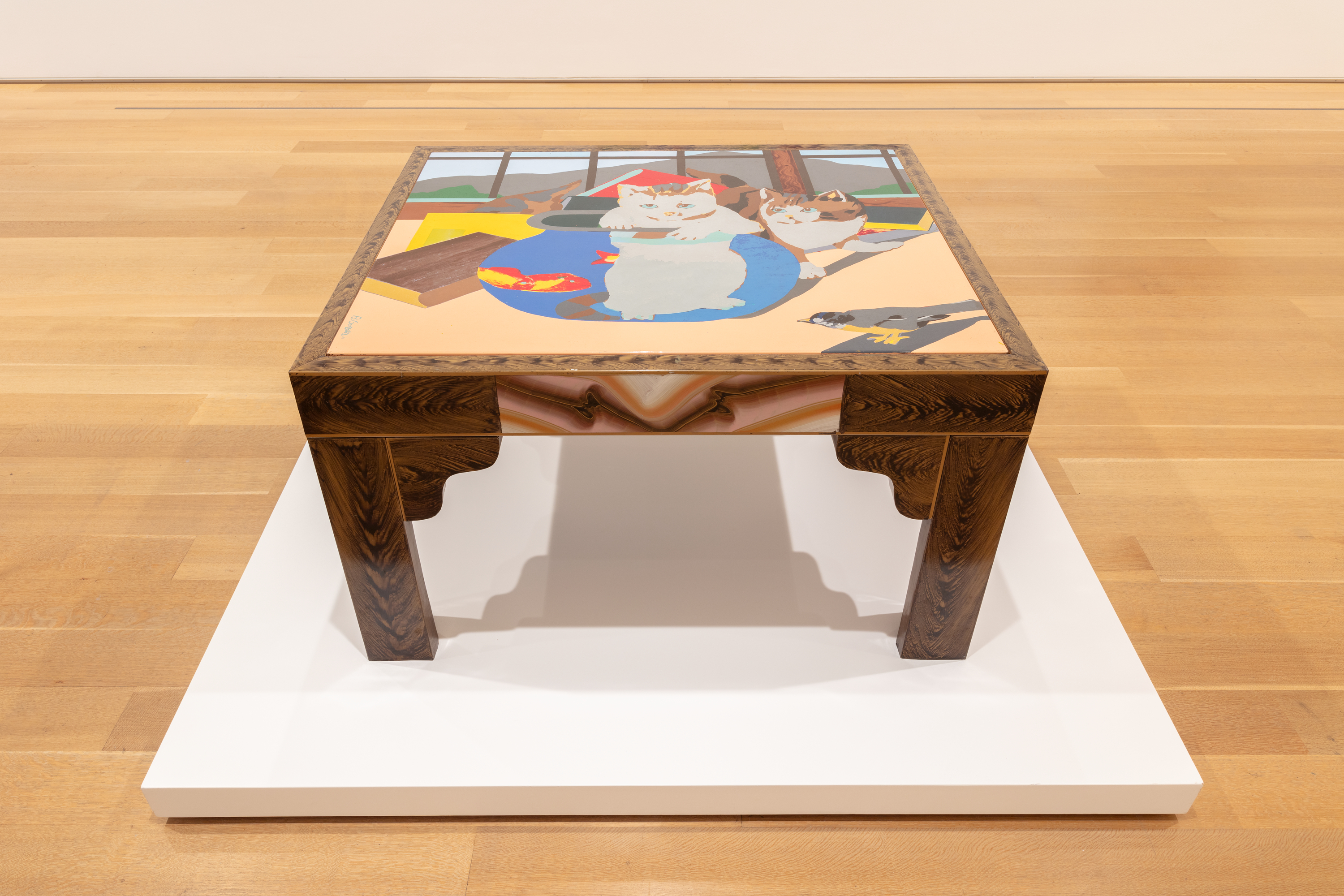 Photo shows a square-topped wooden table featuring the image of many books and two kittens, one in a fishbowl with a live fish.