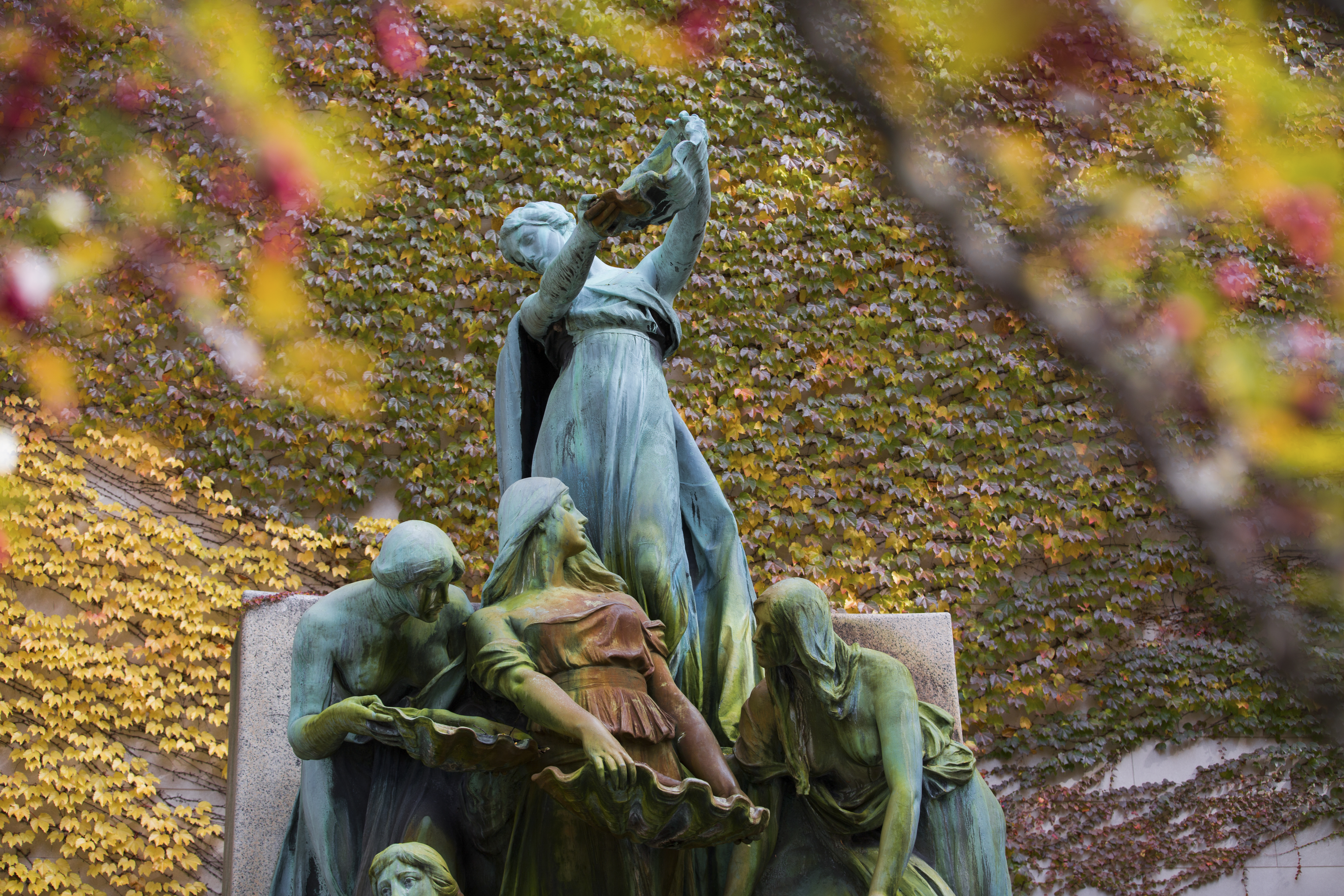 Photograph of the top portion of a bronze fountain, green with patina, featuring several women in draped garments, one prominently at top pouring out a vessel. The fountain is not on, and it is surrounded by green and yellow ivy, the yellow branches of trees blurry in the foreground.