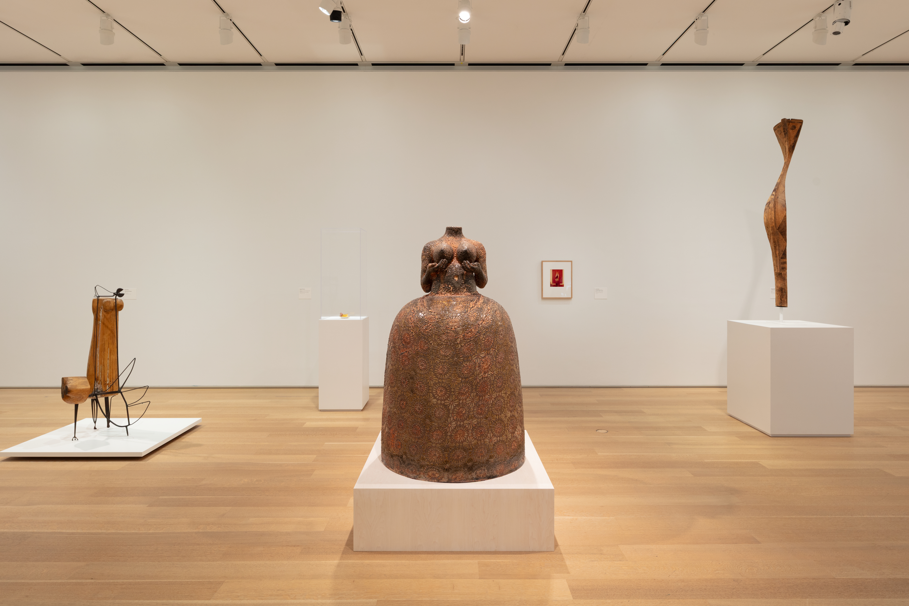 Photo in a white-walled art gallery shows a coppery-brown, life-size sculpture of a headless woman, naked from the waist up, hands nearly cupping the breasts, in a full, bell-shaped skirt. The sculpture is uniformly patterned with an imprinted floral motif.
