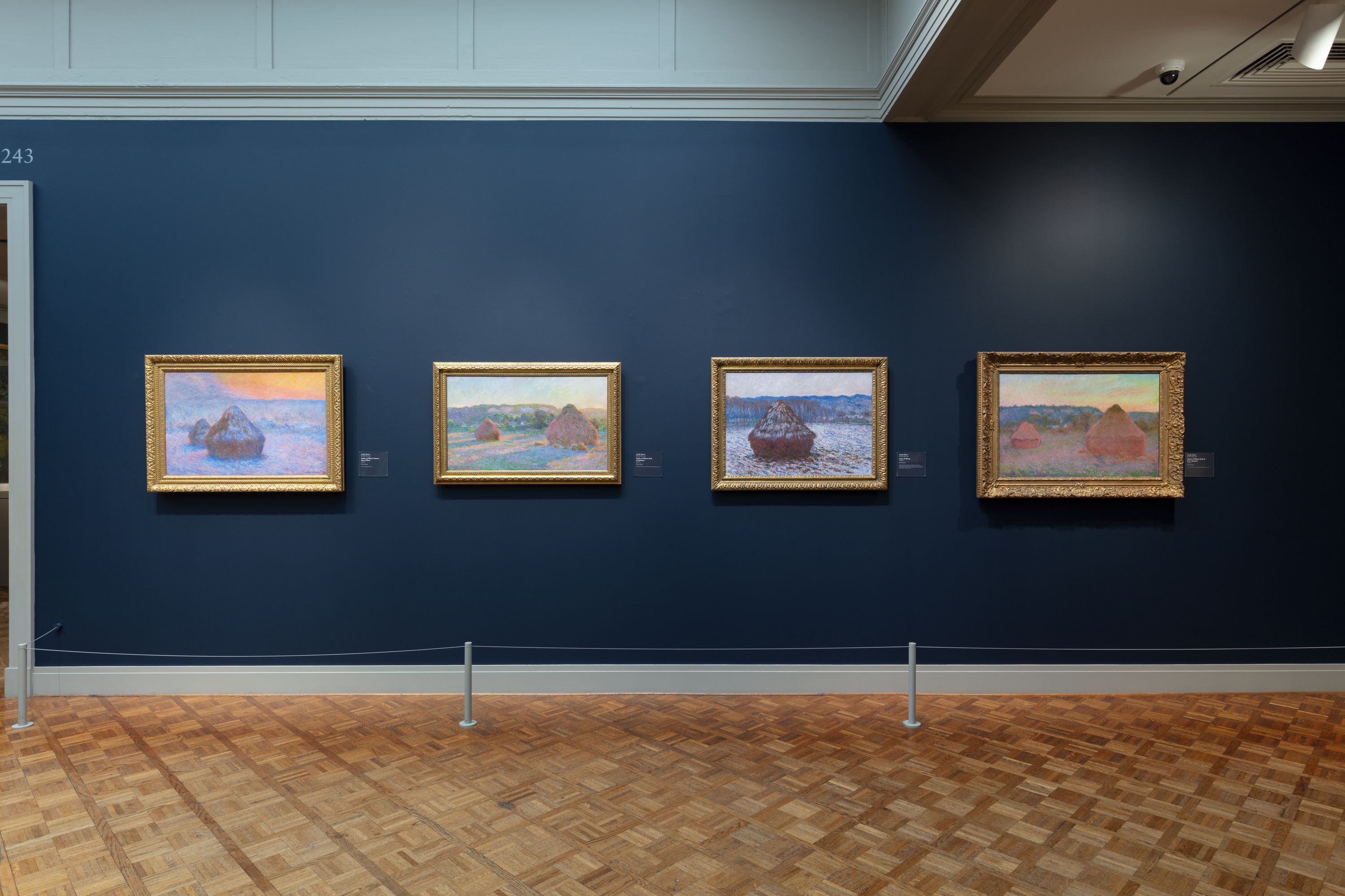 A color photograph shows a museum gallery dominated by four gold-framed haystack paintings on a dark blue wall; at left is a doorway, across the wall the paintings run left to right, below is a light brown parquet floor with gray posts and a rope barrier, and a pale gray ceiling above.