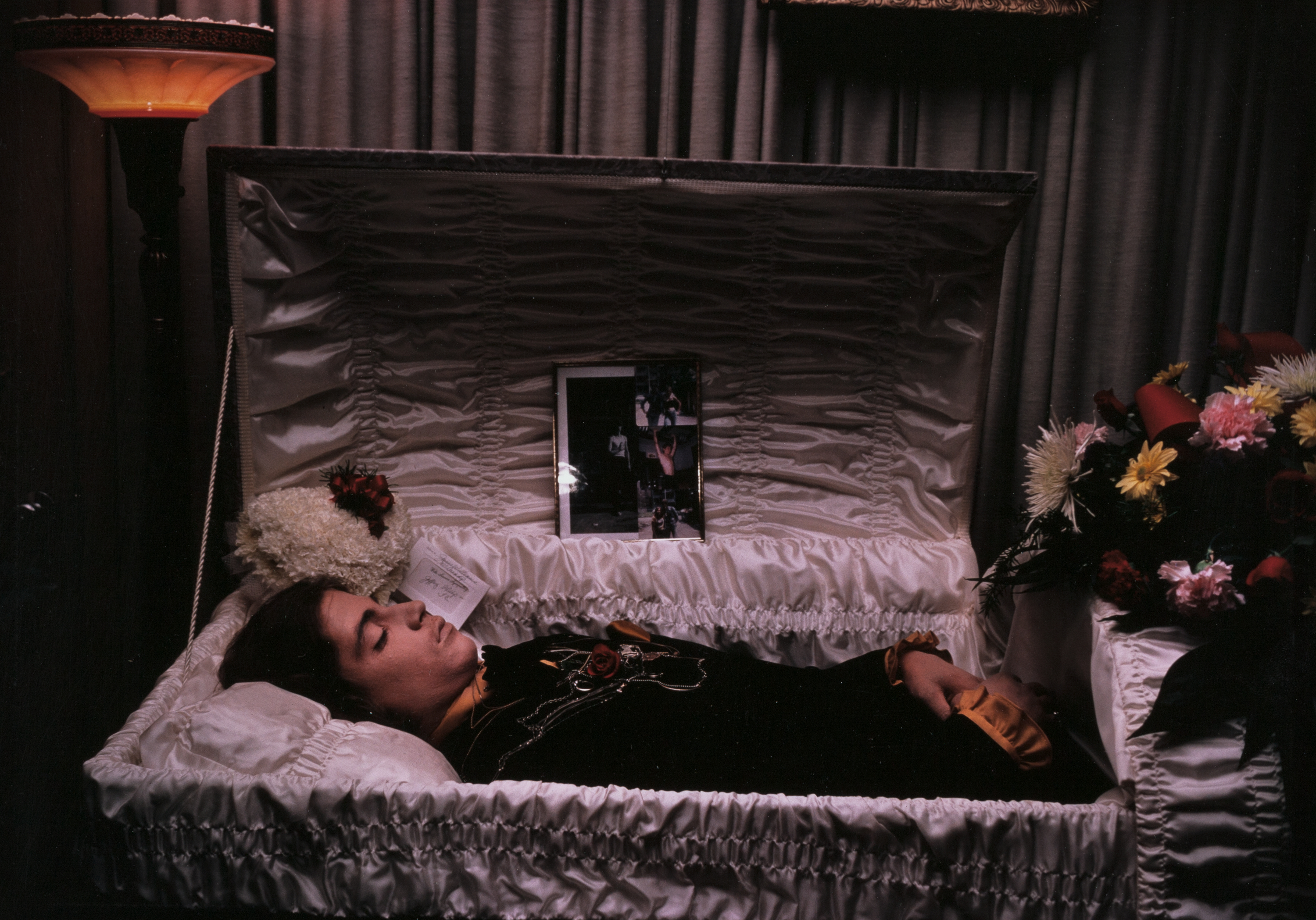 Person with light skin and long dark hair lies in repose inside a white satin-lined open casket, wearing a black garment with gold trim and a red rose pinned to the chest. Their hands are folded across the body. A flower arrangement of white chrysanthemums rests near their head, with a small funeral card tucked beneath. A framed collage of photographs stands upright inside the lid, behind the body. To the right, a large bouquet of pink, red, white, and yellow flowers sits atop the casket edge. A floor lamp and gathered curtains frame the scene.