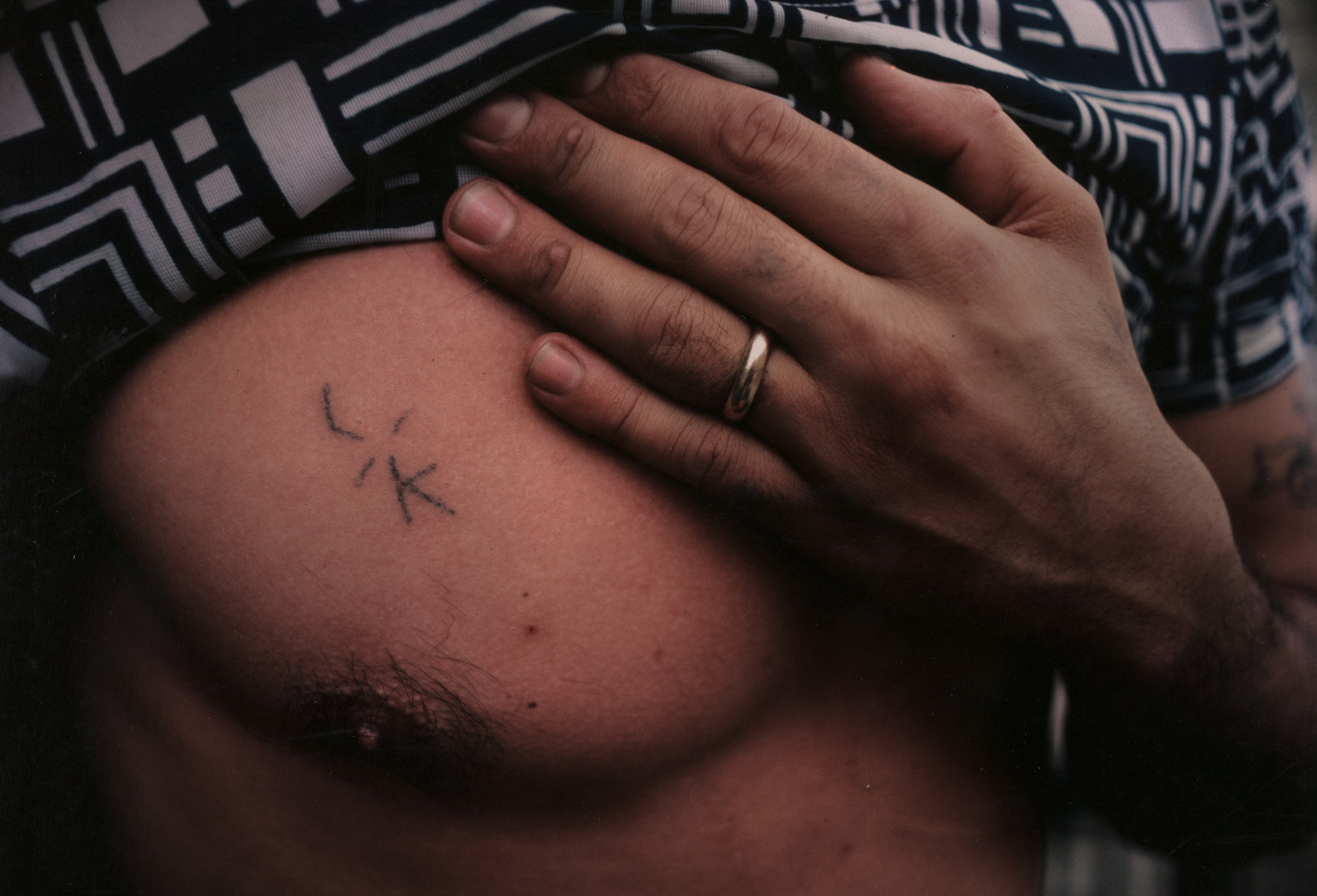Color photograph of a person lifting a patterned shirt to reveal a small, simple black tattoo on their left chest near the nipple. The tattoo appears to form the letters “L” and “K” with short lines radiating outward. A hand with a gold wedding band rests gently on the upper chest, partially obscuring the tattoo. The image focuses on skin texture, hair, and the intimacy of the gesture.