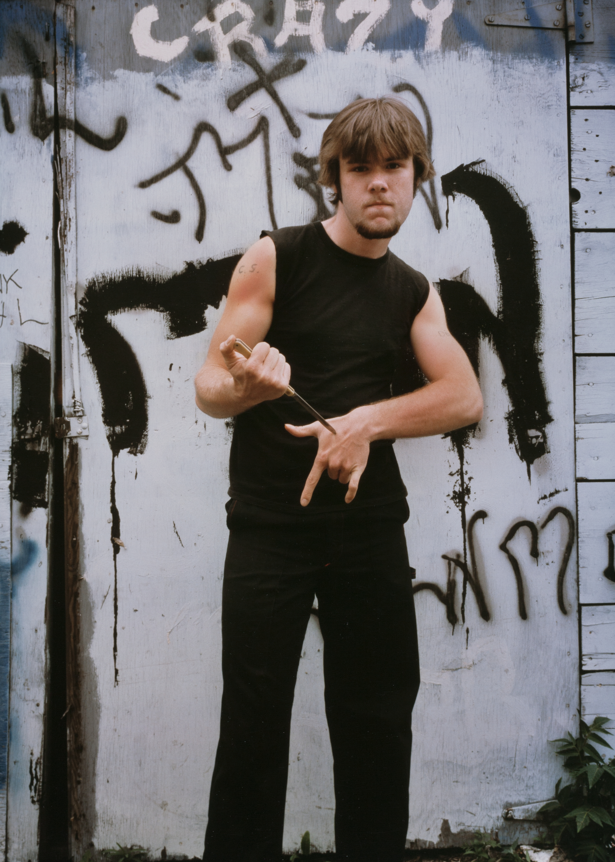 Young man standing in front of a graffiti-covered wooden wall. He wears a black sleeveless shirt and black pants, holding a metal rod or knife horizontally while gesturing with his other hand. The graffiti includes bold black and white spray-painted shapes and the word “CRAZY” written above his head in large white letters. His stance is assertive, and he looks directly at the camera.
