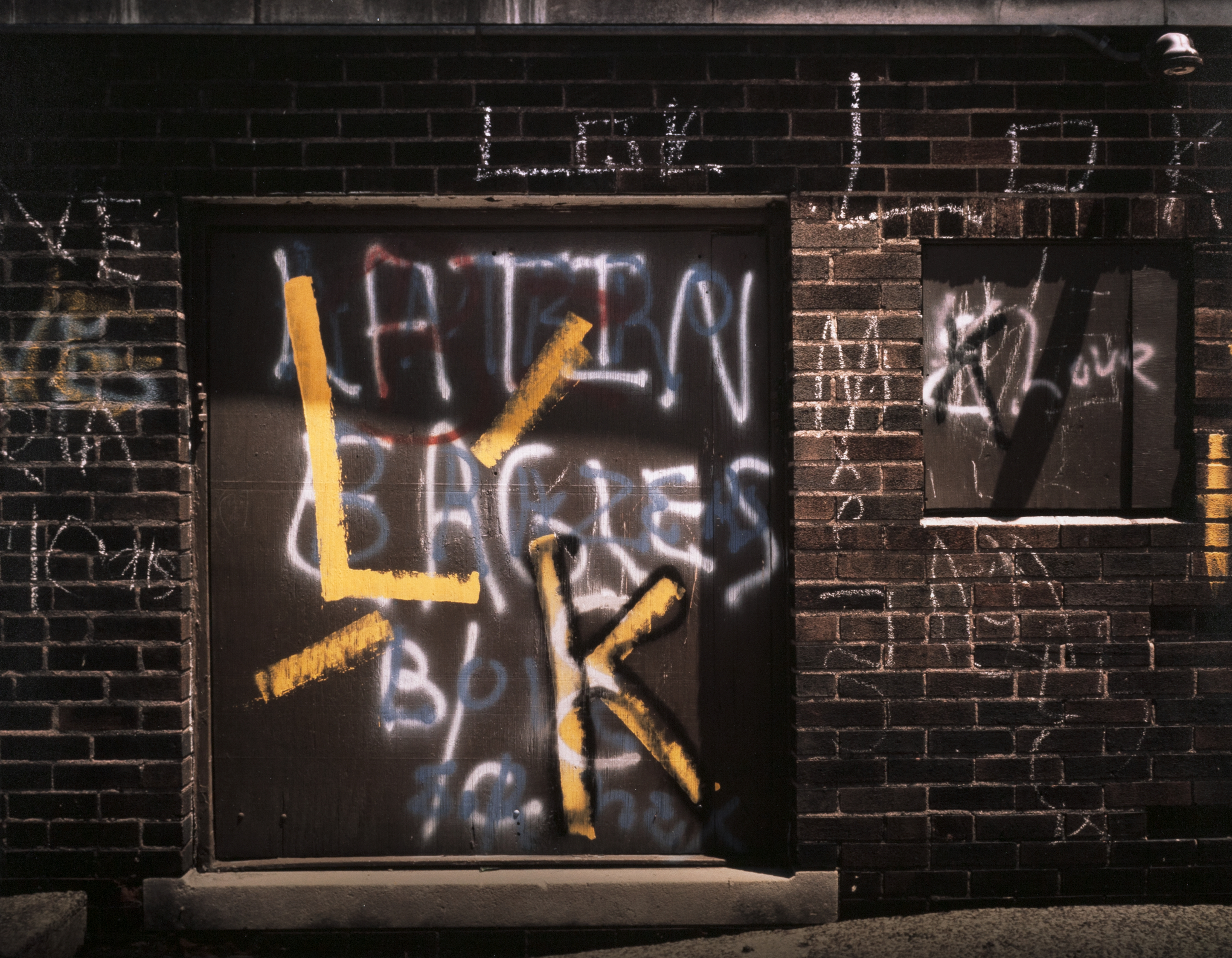Graffiti-covered brick wall with a boarded-up doorway and window. The door is painted with large overlapping spray-painted letters and symbols in yellow, white, blue, and red. Prominently, a large yellow “L” intersects a yellow “K” over white spray-painted text that includes “LATIN BLACKS” and “B.O.C.” The surrounding bricks are marked with chalk and spray paint, including more “L” and “K” initials, crossed-out symbols, and partially legible names or words. A small security camera is mounted above the window at upper right.