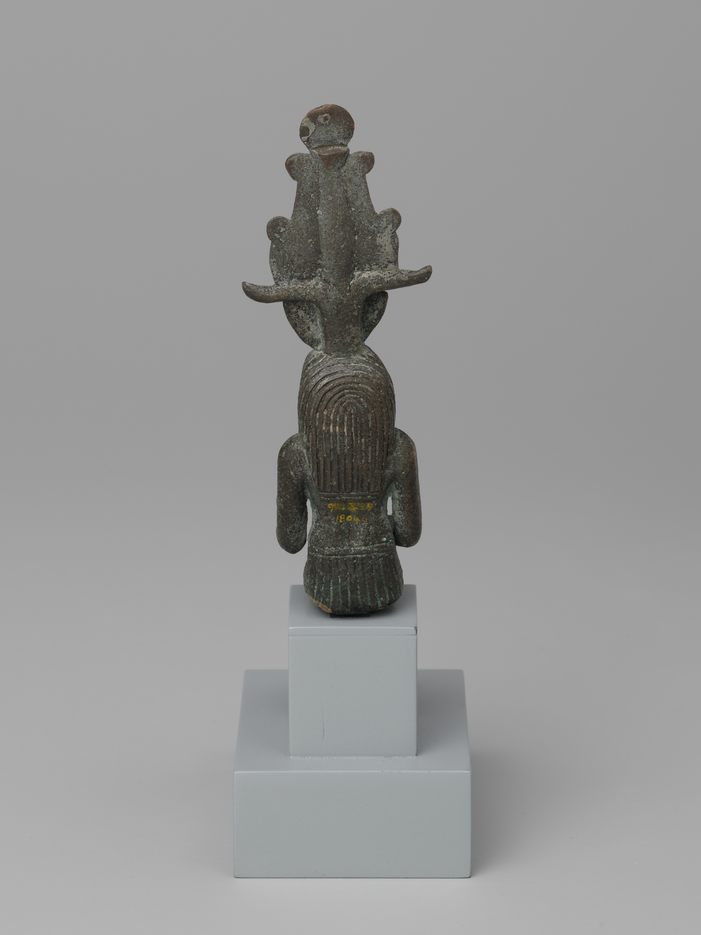 At the top, a small rounded piece with two pale circular marks sits above a tall, branching headdress in dark green-brown. Below, a human-like figure is shown from the back, with a head covered in tight vertical ridges, short arms hanging at the sides, and a ridged lower section reaching about mid-thigh. Centered on a light gray two-tier pedestal, the figure appears small relative to the stand, with plain gray space surrounding it.