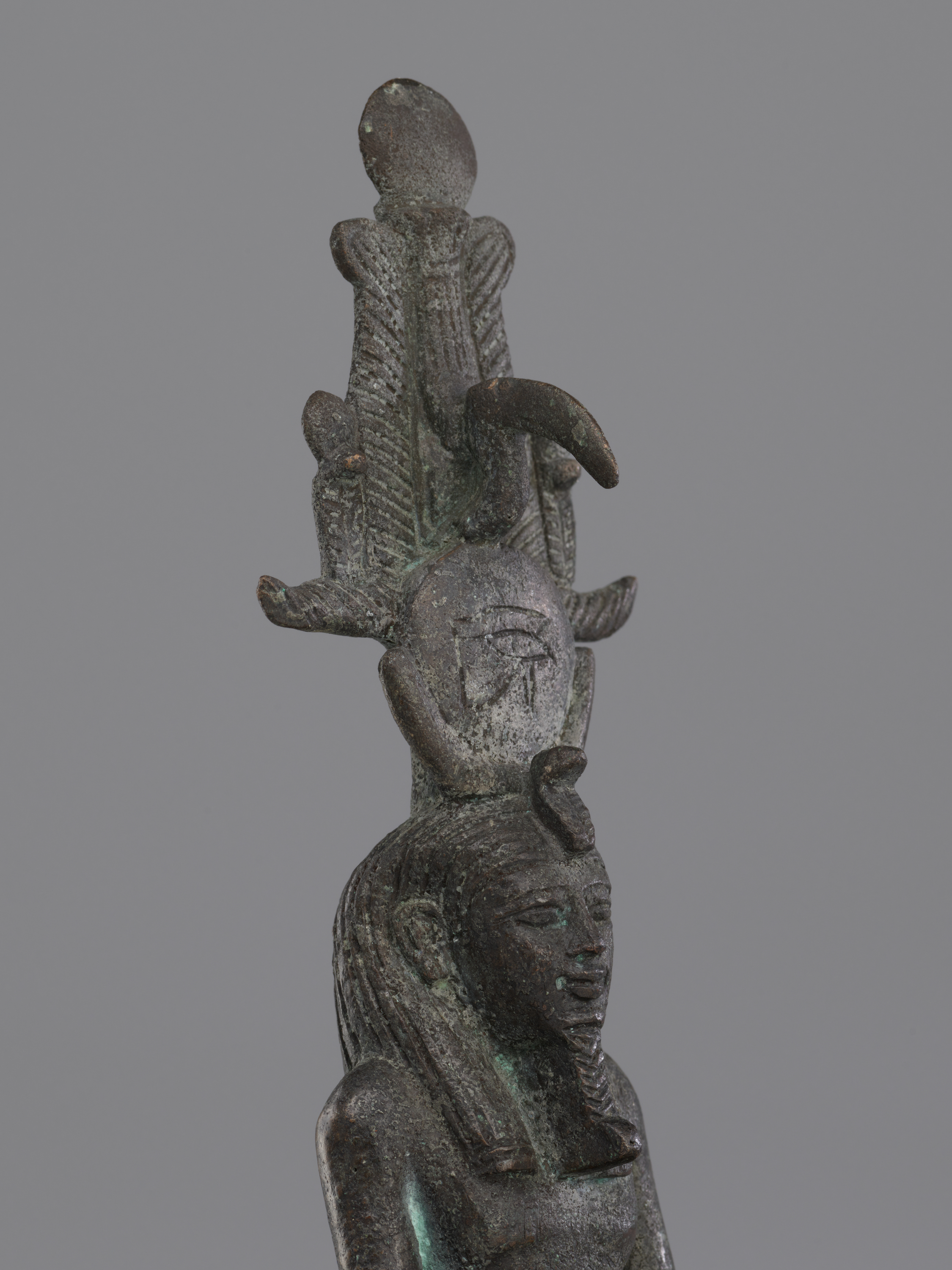 Top to bottom: a rounded disk sits above three tall, textured vertical shapes, with two short curved horn-like pieces projecting from the sides. Beneath is a circular head; moving downward, long ridged strands drape over the shoulders and a narrow torso continues with arms held close to the sides. The colors are dark gray with small areas of green, and the headdress is taller than the head, giving the figure a slender, upright appearance in a single scene.