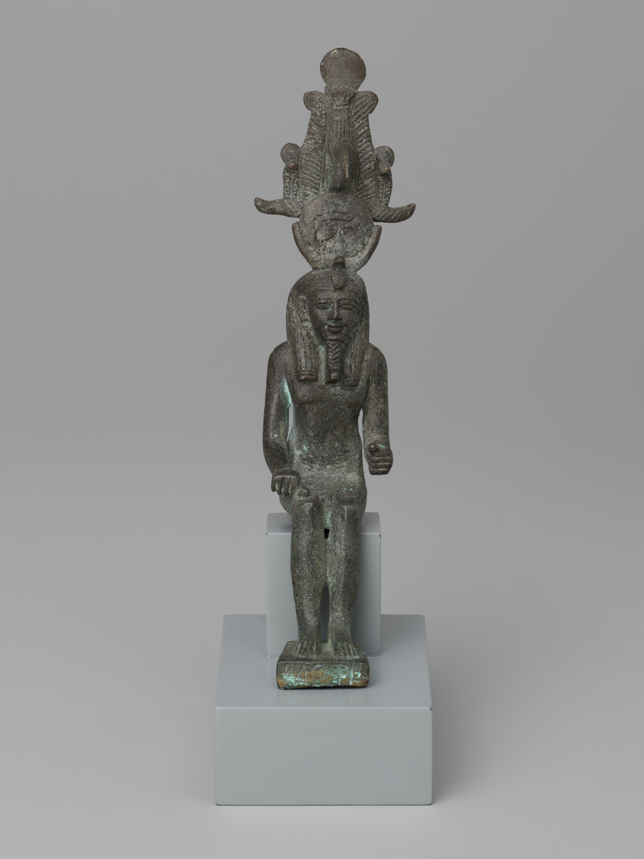 From top to bottom, a dark green-gray standing figure wears a tall crown with two vertical plume-like shapes and a round disk, centered against a light gray background. In the middle, the front-facing figure has a long neck with a ridged collar and straight arms held close to the sides. At the bottom, straight legs end in flat feet on a small rectangular base with hints of blue-green, which rests on a larger two-tier light gray pedestal, making the figure appear small relative to its stand.