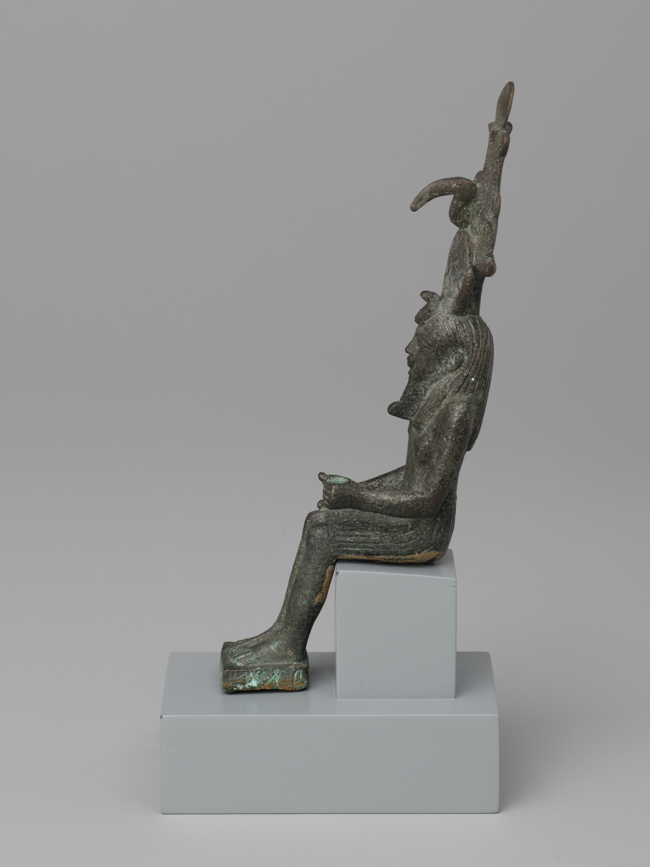 At the top, a tall vertical ornament rises from the head with several curved projections. Below, a seated human-like figure in left profile has a pronounced nose, a ridged hair or head covering, and arms bent with hands resting together on the lap. At the bottom, bent legs extend forward to flat feet on a small rectangular base; the figure is dark green-brown with lighter green areas and is mounted on two stacked light gray blocks against a plain light gray background.