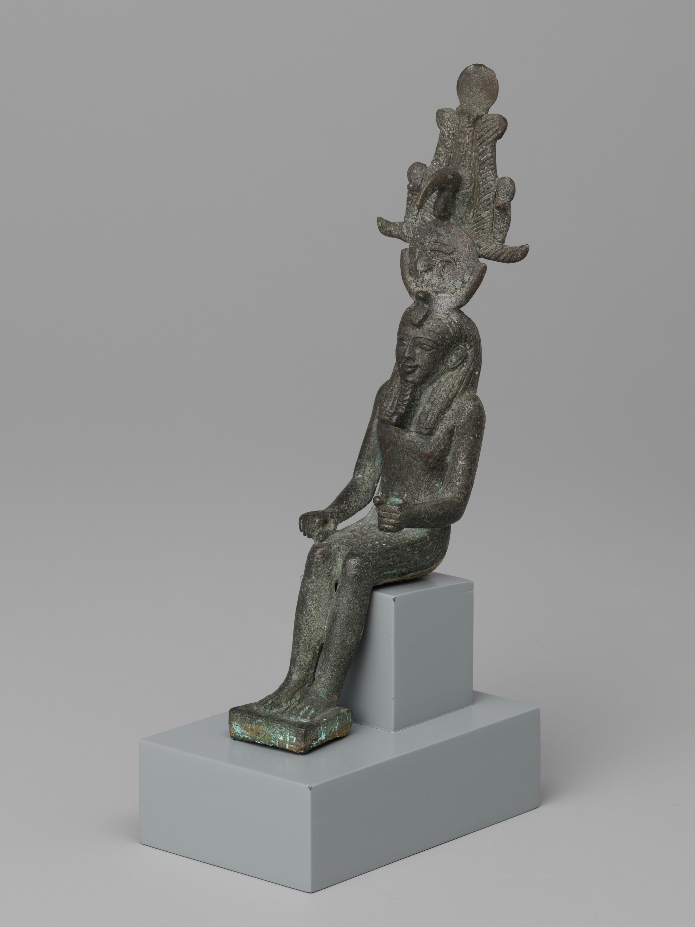Top to bottom: a small dark green seated human figure wears a tall, ornate headpiece that rises to a round element at the top. The torso is upright and the arms are bent, with hands near the lap; shallow ridges are visible across the chest and body. Legs extend forward with flat feet, and the figure sits on a small rectangular block placed on a larger light gray stepped base, facing toward the right against a plain light gray background.
