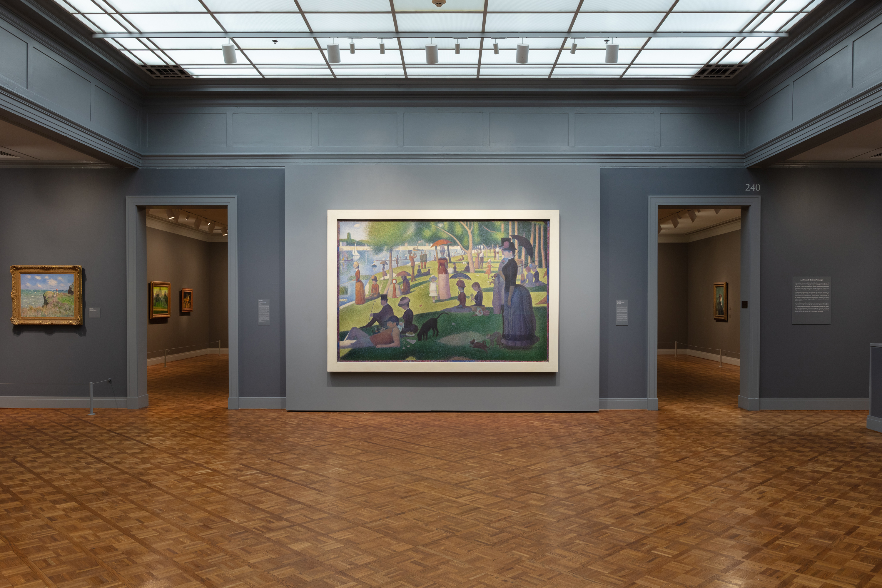 Photograph shows Georges Seurat’s large painting "A Sunday on La Grande Jatte—1884" hanging on a blue-gray wall between two open doorways to other galleries wearing a flat, wide, sharp-cornered white frame.
