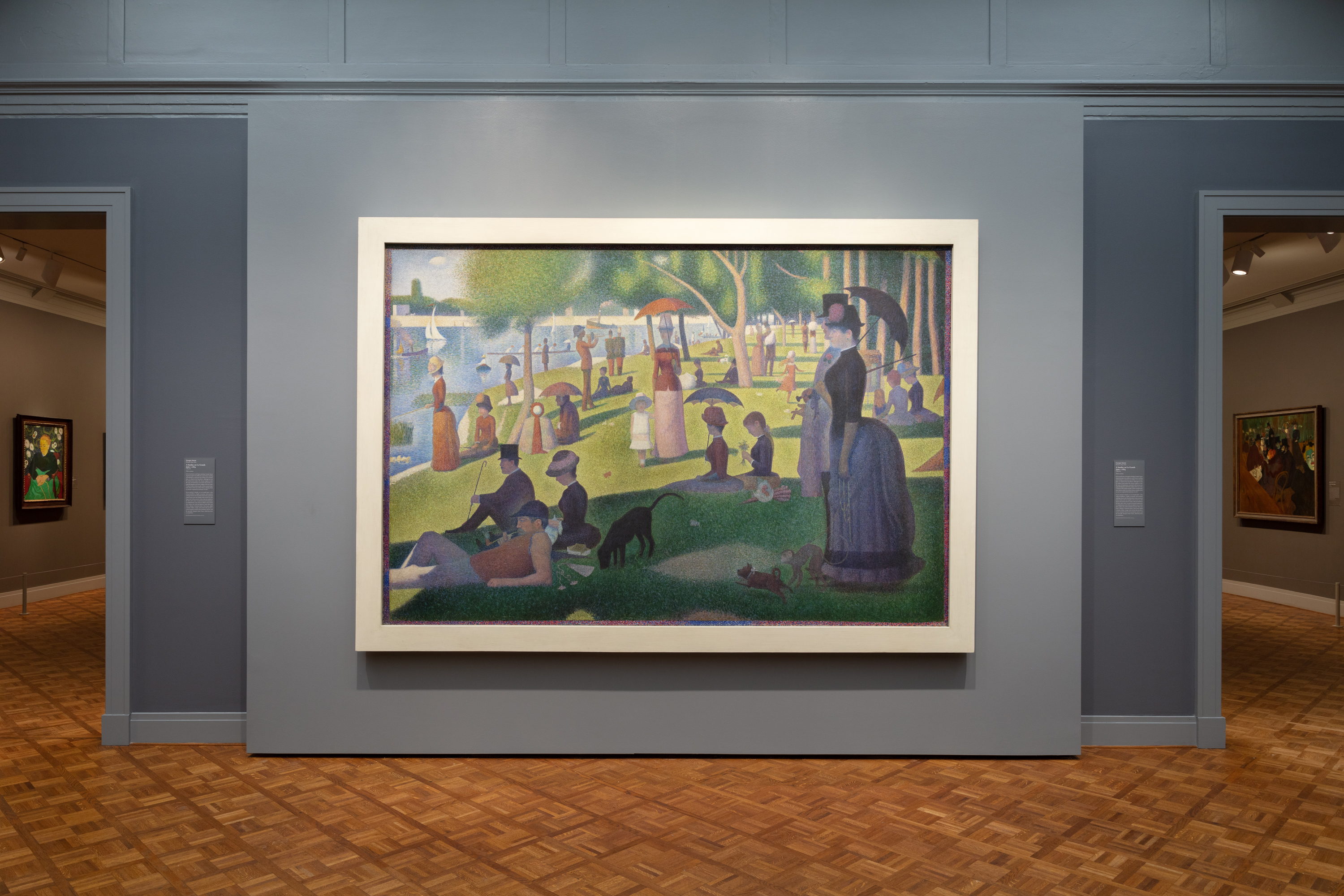 Photograph shows Georges Seurat’s large painting "A Sunday on La Grande Jatte—1884" hanging on a blue-gray wall between two open doorways to other galleries wearing a flat, wide, sharp-cornered white frame.