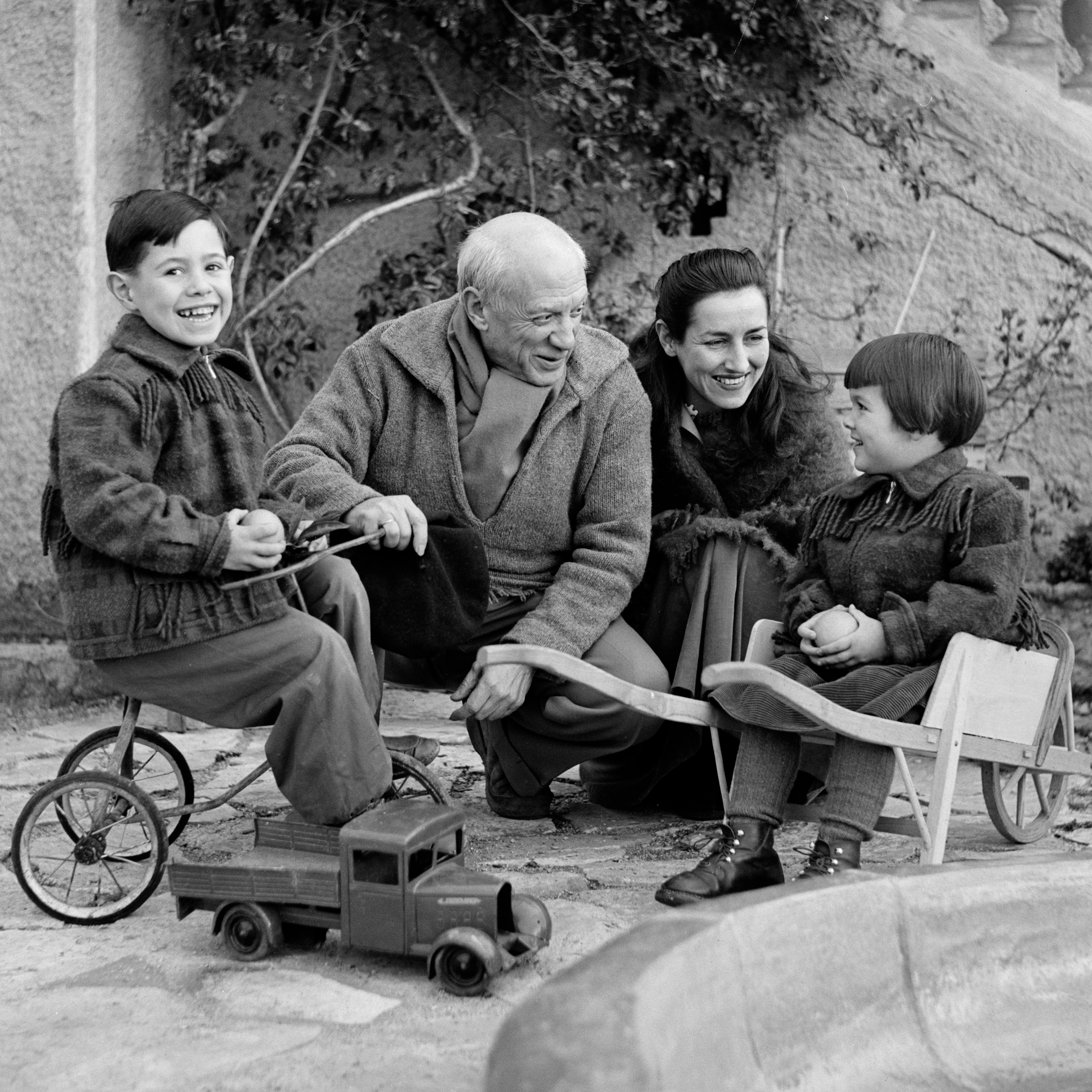 A black-and-white photo shows the artist crouching next to his partner, Françoise Gilot, and smiling at their 4-year-old daughter, Paloma, who sits in a small wheelbarrow smiling back at her parents while their 6-year-old son, Claude, sits on a tricycle on the other side of father, smiling at the camera.