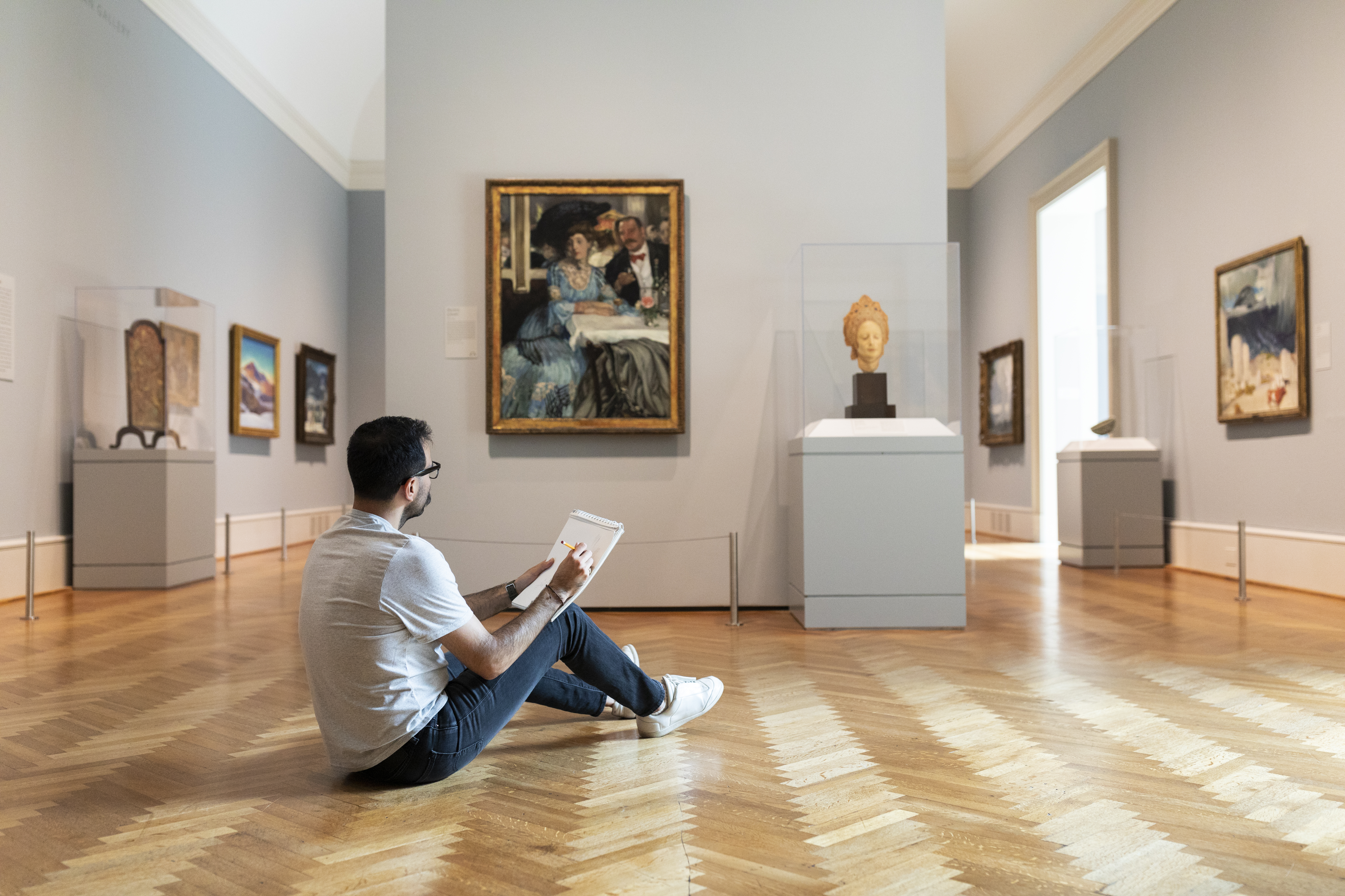 Photograph of a young light-skinned man with short black hair sitting on the floor of an art gallery holding a sketchpad, his back partially toward the viewer,  as he looks toward a painting of a woman in blue.