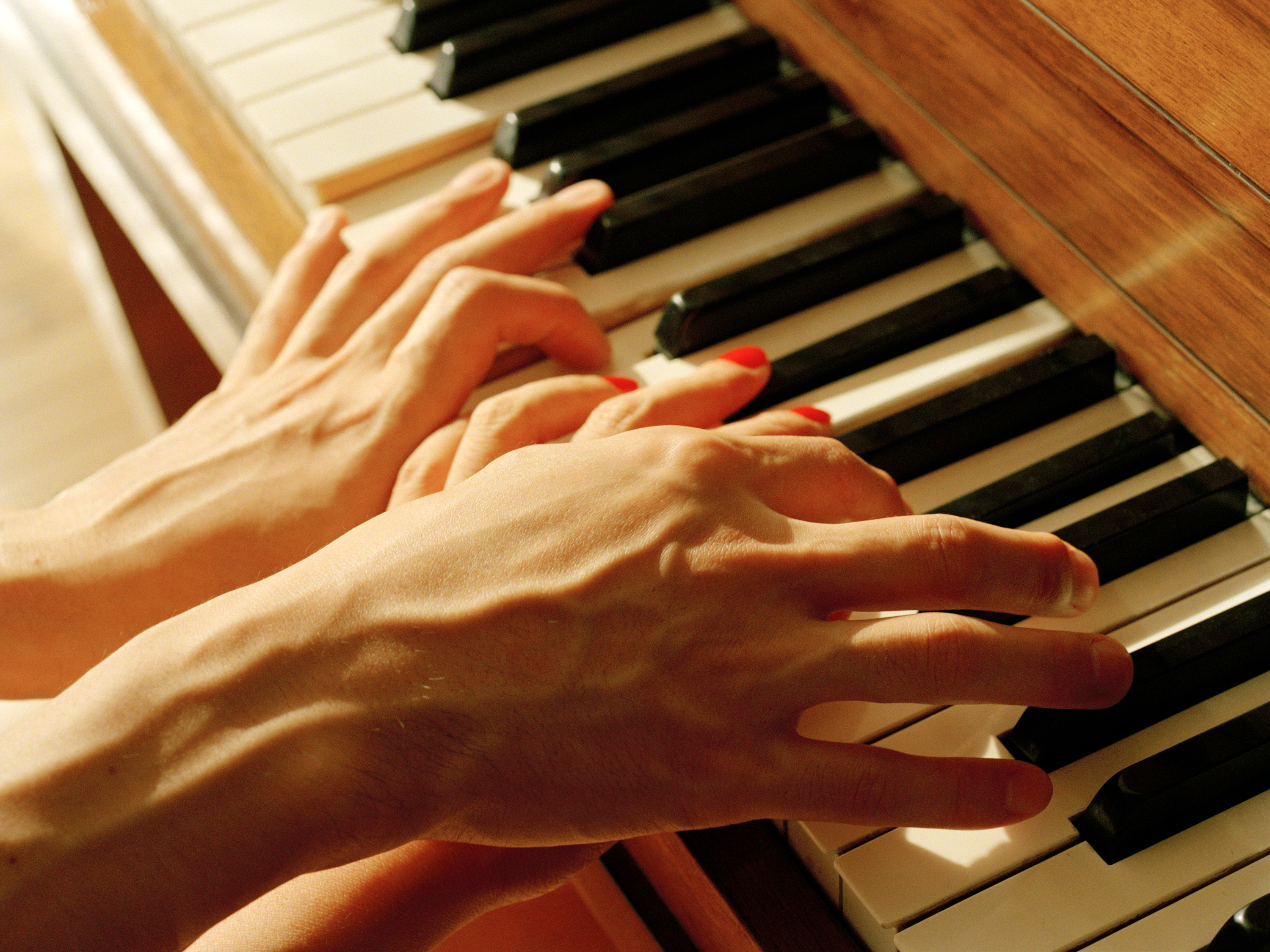A color photograph of two sets of light-skinned hands, intertwined as they play the piano. One set has red nail polish; the other none.