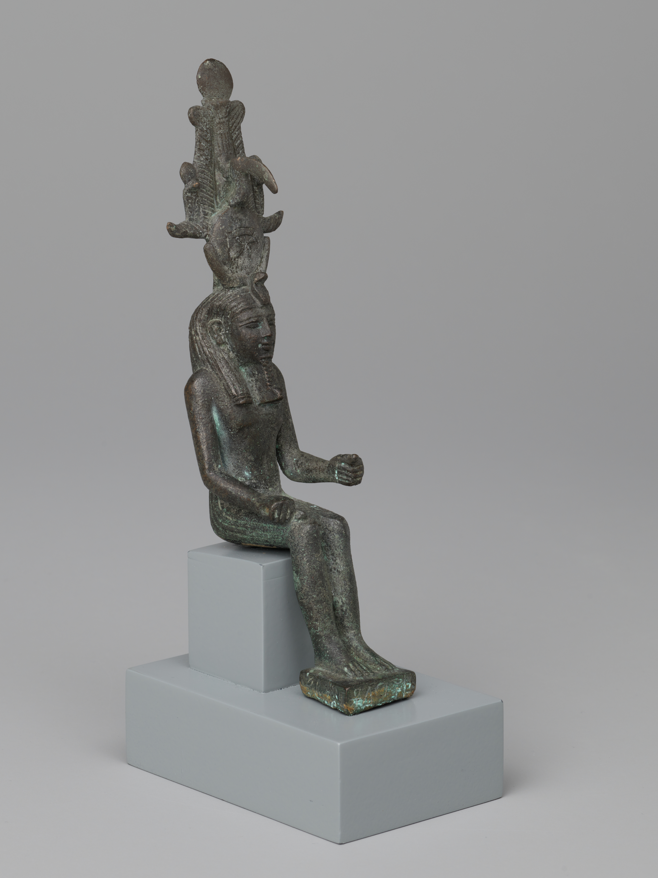 Top to bottom: a seated human figure faces toward the left, wearing a tall, tiered headpiece with a raised snake form and curved elements projecting outward. Moving down, the torso is upright with the right arm bent forward in a closed hand, while the left arm rests along the side; the surface is dark green-brown with lighter green areas. At the bottom, the legs extend together to a flat footrest, and the figure sits on a small rectangular seat placed on a larger, light gray stepped block, making the figure appear small relative to its base.