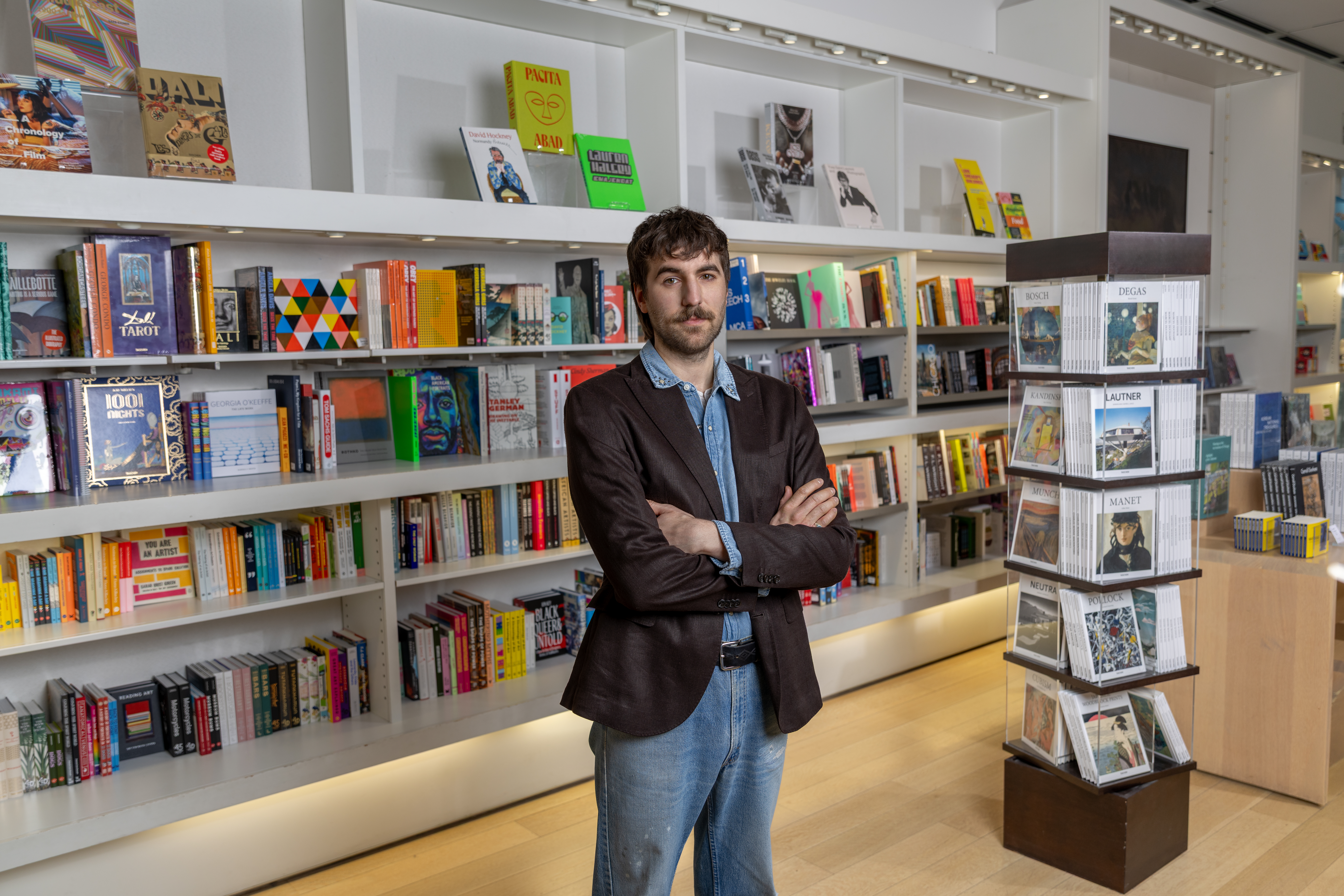A color photograph of a bookstore interior shows at center an adult with short dark hair and light skin wearing a brown blazer, blue shirt, and jeans with arms crossed, with long white shelves of colorful books at left and behind, and a tall rotating display rack at right.