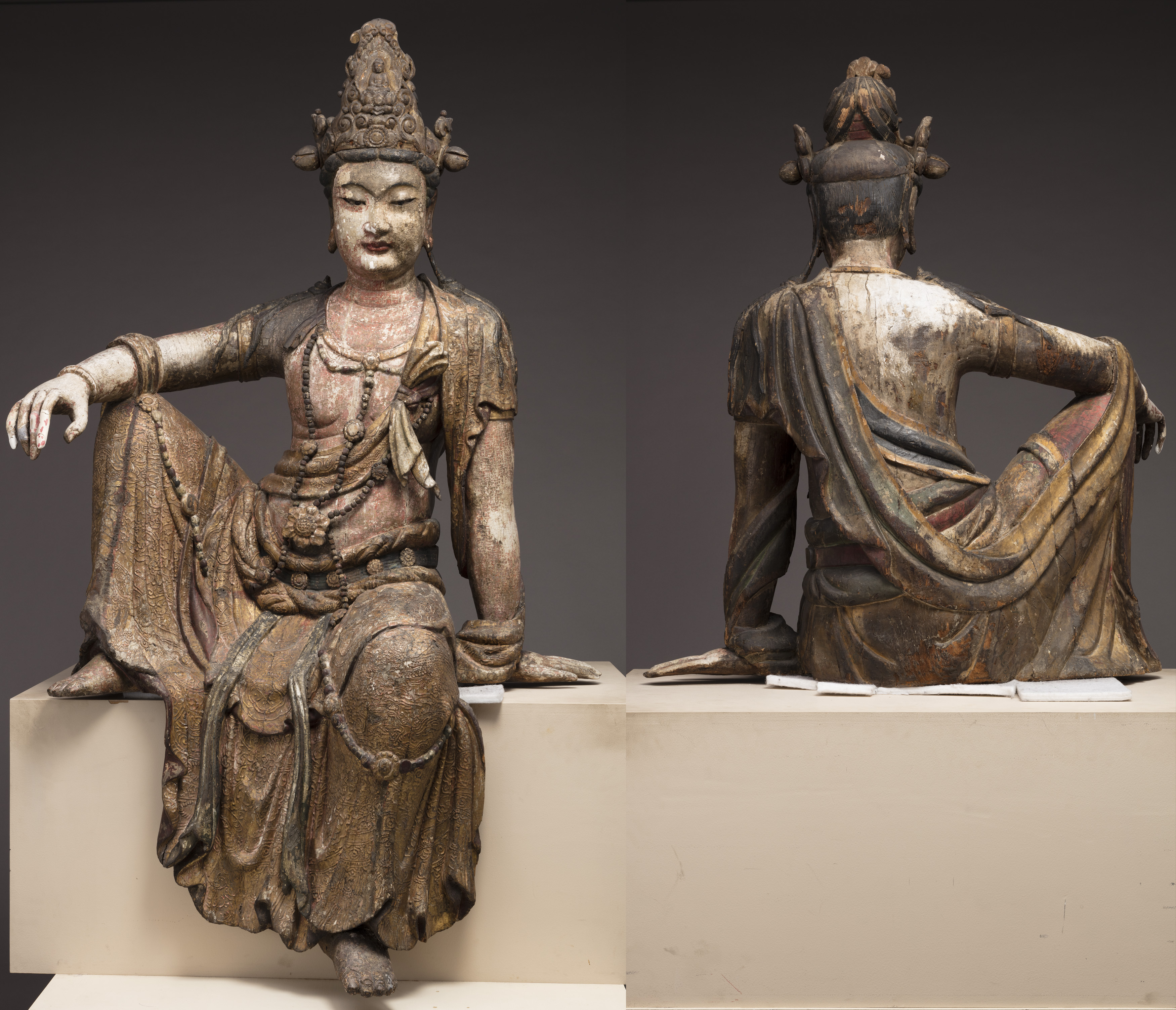 Front and back views of a carved statue of a figure seated jauntily, his left leg bent up on a platform. The surface is mottled and colored in places, pale beigey-pink or brown in others. He wears carved robes, a belt, and an elaborate headdress. His eyes are wide-set and partially closed, his hair in braids.