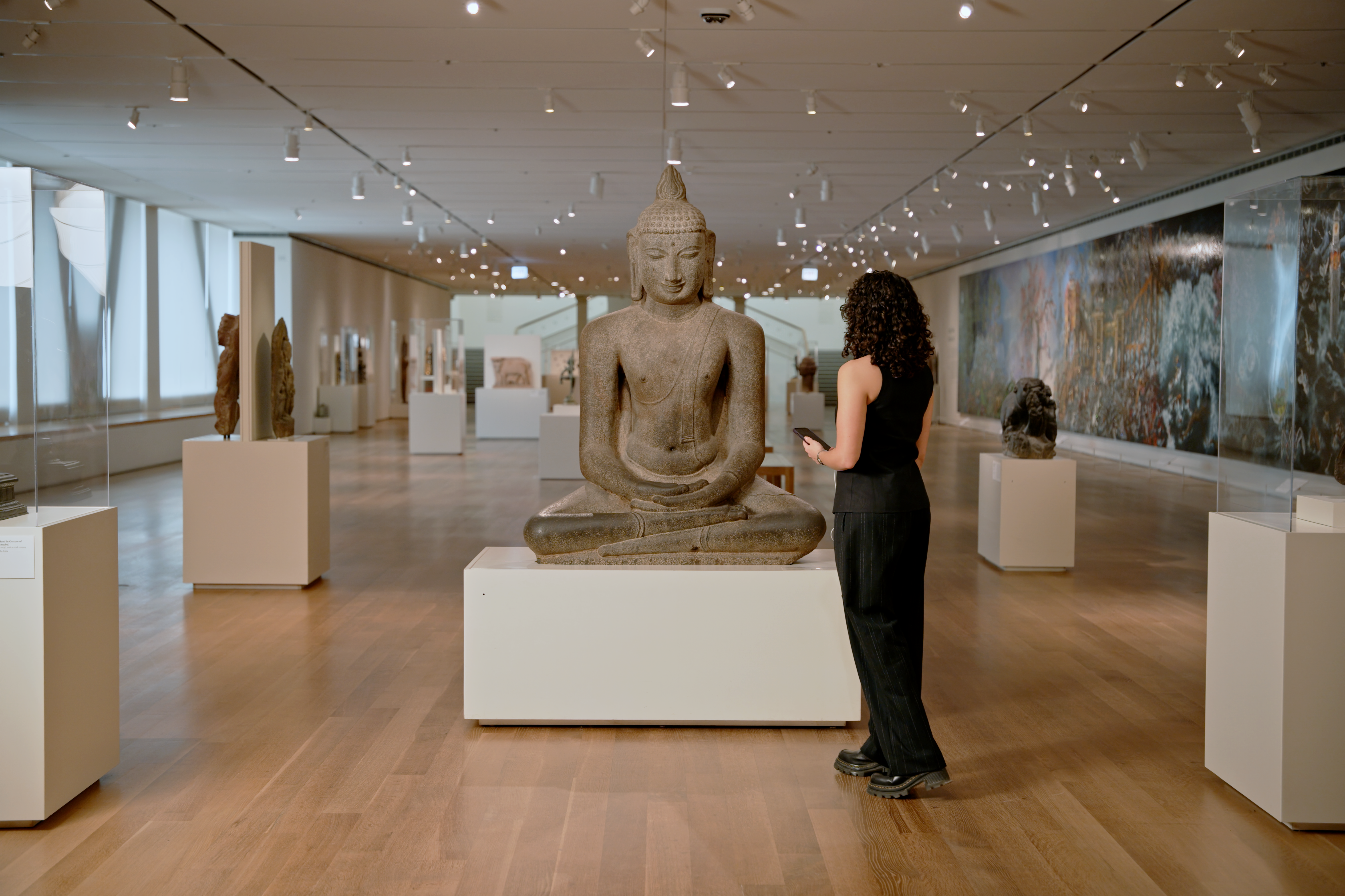 Color photograph of a museum gallery with wood floors, centered on a large gray stone seated figure on a white pedestal; at right an adult with curly dark hair in black clothes stands, with tall windows and pedestals at left and a long mural and more sculptures along the right and background.