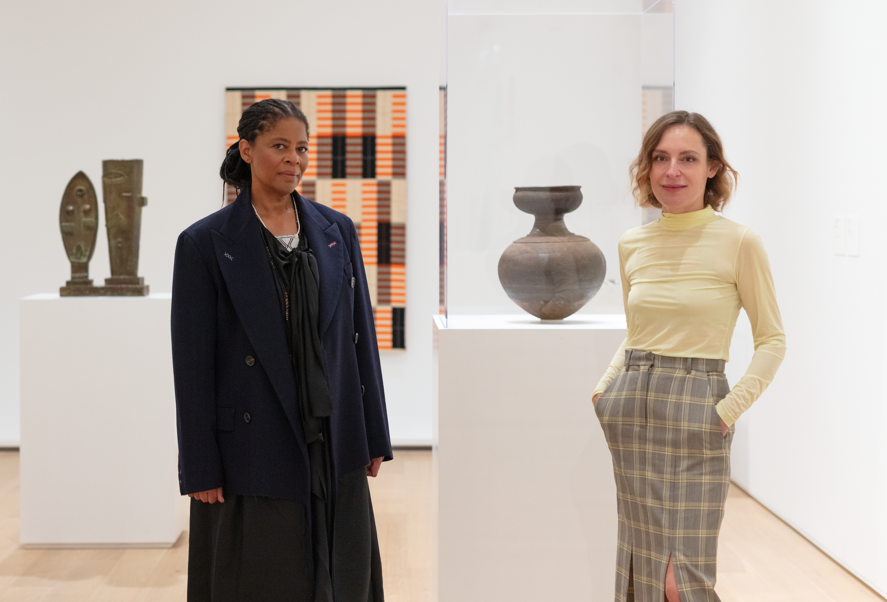 Two women, Simone Leigh—a Black woman with braids in a structured navy sportcoat—and Paulina Pochoba—a light-skinned woman with short, wavy highlights and a yellow turtleneck—stand together in a white gallery space, facing the viewer. Behind them, from left to right, is a bronze-colored sculpture in a vitrine, a red and black textile, and a ceramic pot.