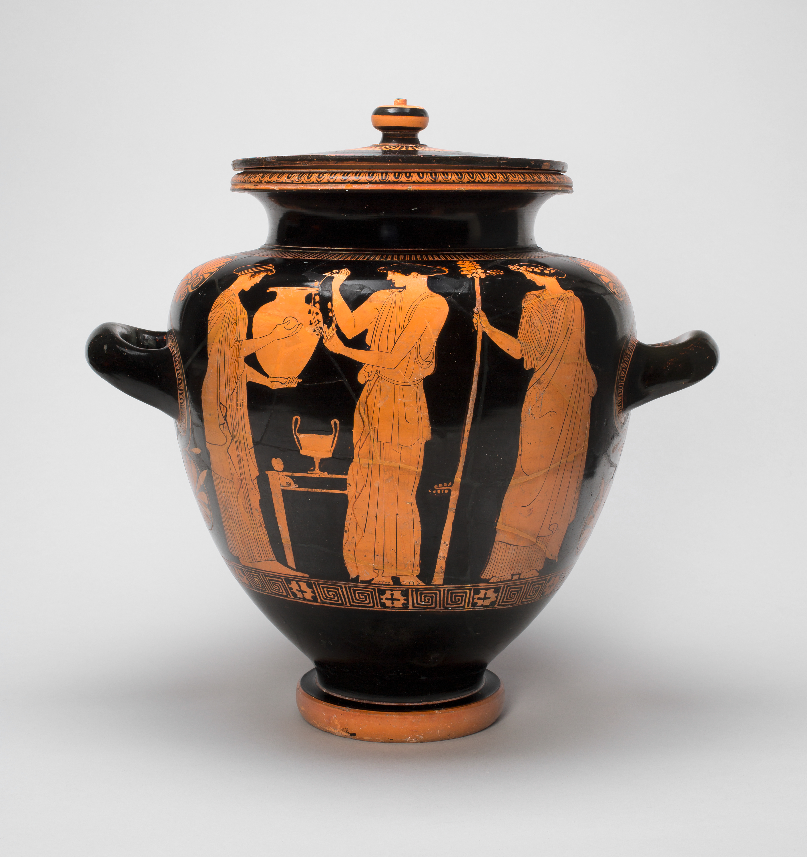 A dark-colored jar featuring, in orange on one side, a female figure in robes holding a large vessel out toward another robed figure, who reaches to place an object, maybe a necklace, inside it. A third figure, at right, holds a staff.