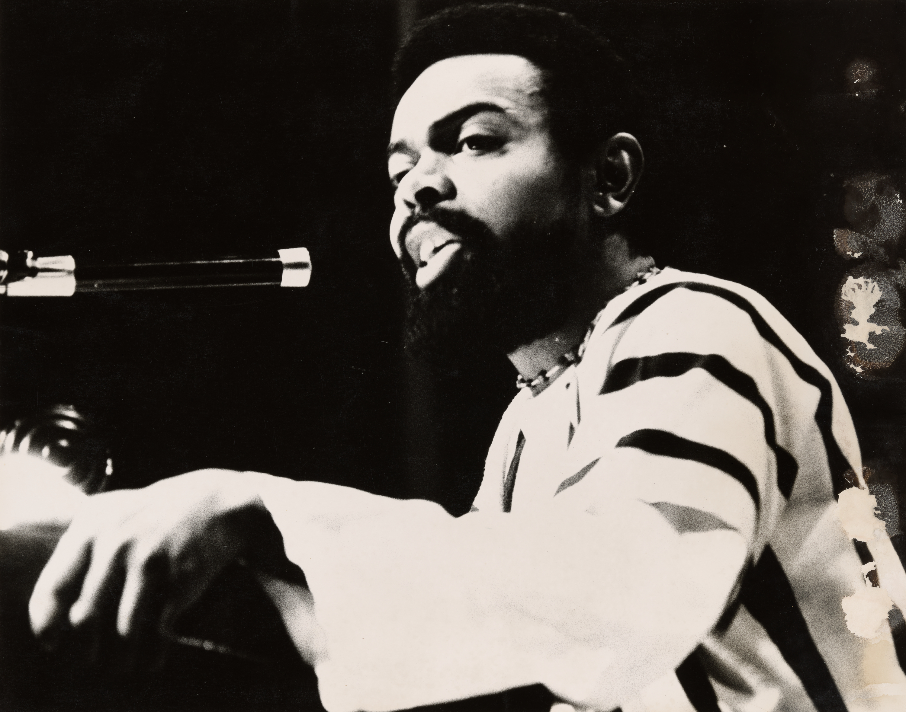 Black-and-white photograph of a man speaking or performing into a microphone. He has a short afro and full beard, and wears a striped garment with wide sleeves and a beaded necklace. His face is lit from the front, while the background is dark, drawing attention to his expression and gestures. His posture suggests focus and intensity, with one hand raised near the microphone.