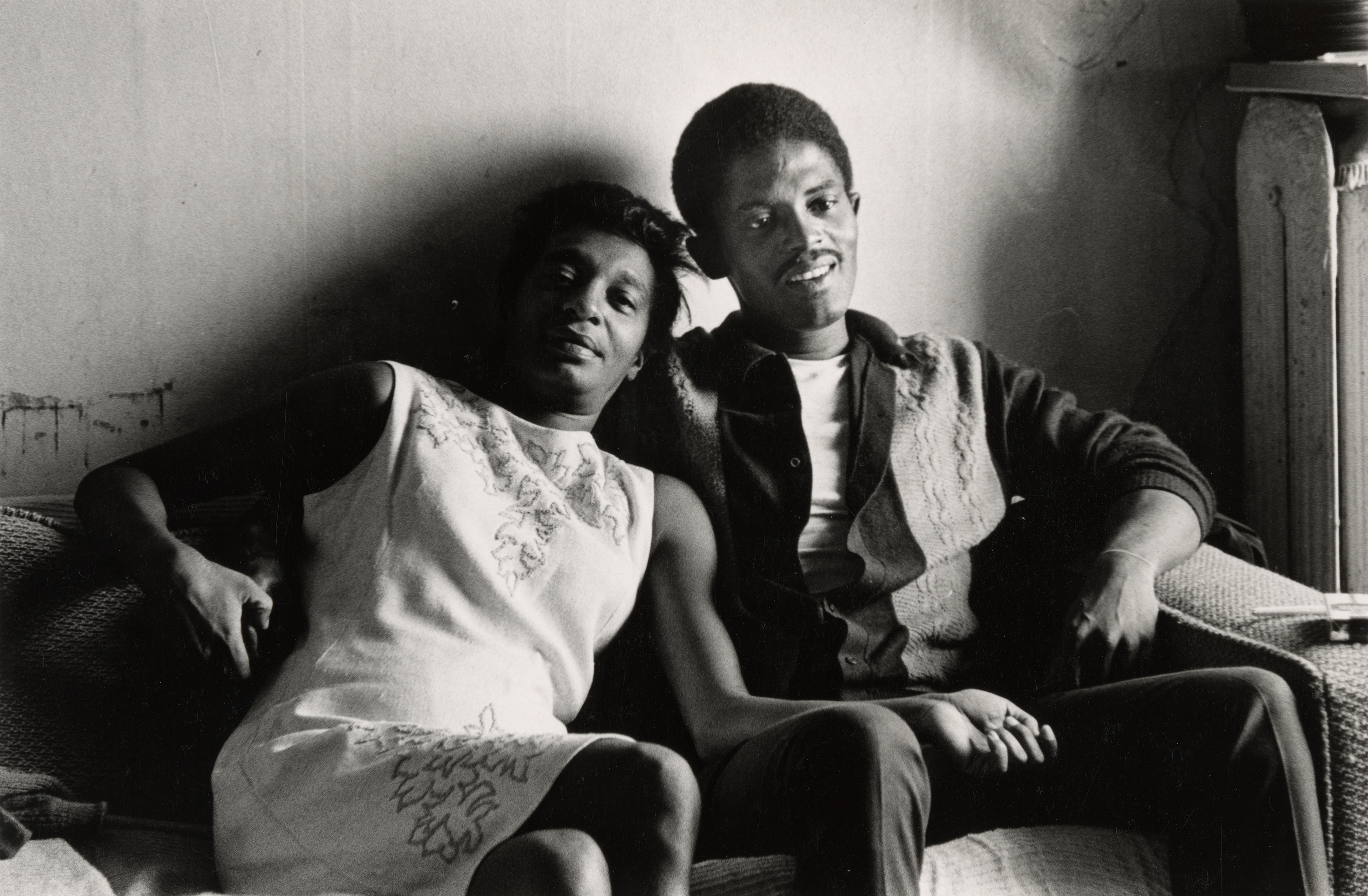 Black-and-white photograph of a couple sitting close together on a couch in a dimly lit room. The woman on the left leans into the man, resting against his shoulder, and wears a light-colored sleeveless dress with floral embroidery. The man, seated on the right, wears a dark shirt under a light cardigan and has one arm around her and the other resting on his leg. Both look relaxed, with soft expressions. The wall behind them is worn, and the corner of a table or mantle appears at the edge of the frame.