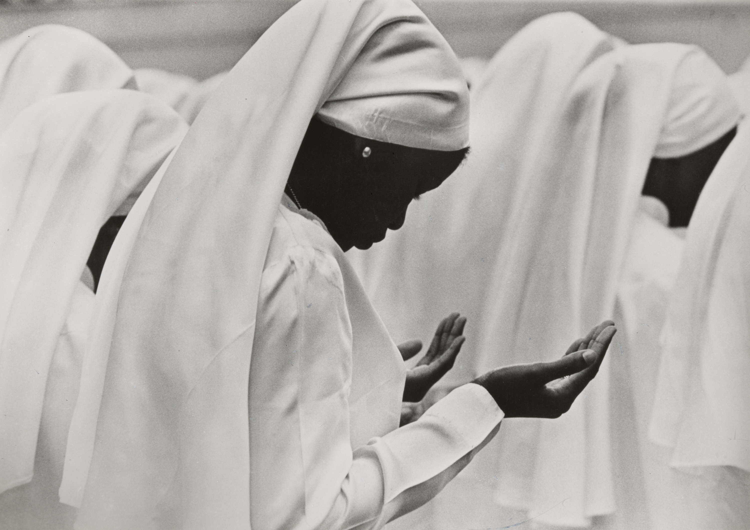 Black-and-white photograph of a group of women wearing white garments and head coverings, standing in prayer. The central figure is shown in profile with her head bowed and palms open in front of her chest. She wears a pearl earring and stands out against the soft, flowing white fabric of the others around her. The composition emphasizes stillness and reverence, with a focus on the quiet intensity of the prayerful moment.