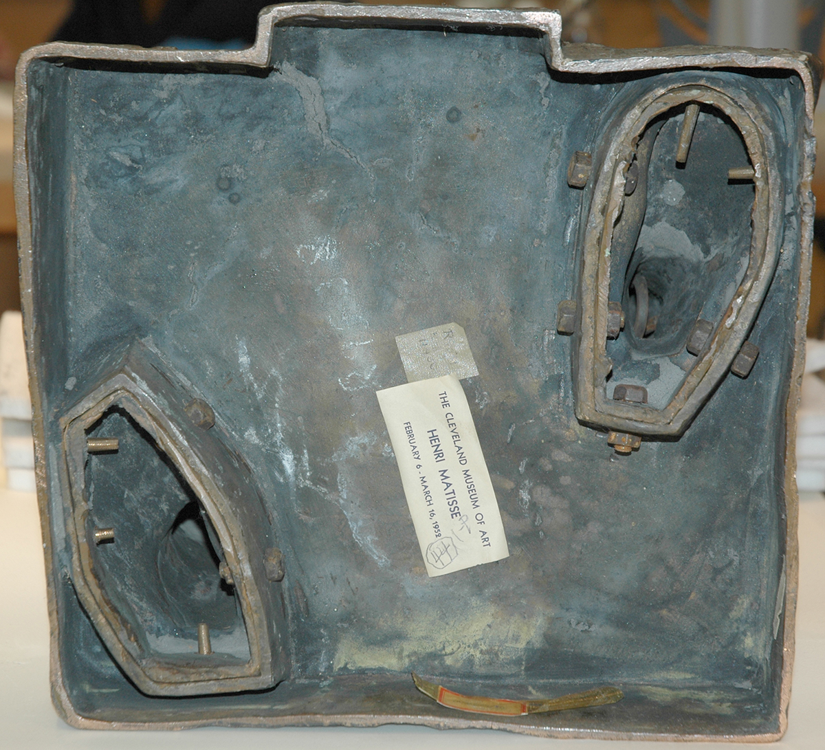 At the top edge is a shallow rectangular notch, with a gray-blue surface and brown rim surrounding the interior. Moving down, an oval recessed section with raised edging sits on the upper right, containing several short cylindrical pegs and a hollow center; a smaller, similarly shaped recess appears on the lower left. Near the center, a cream paper label affixed at a diagonal shows printed text including “The Cleveland Museum of Art” and “Henri Matisse,” with a small piece of tape above it. Along the bottom edge, a narrow curved strip rests near the center.