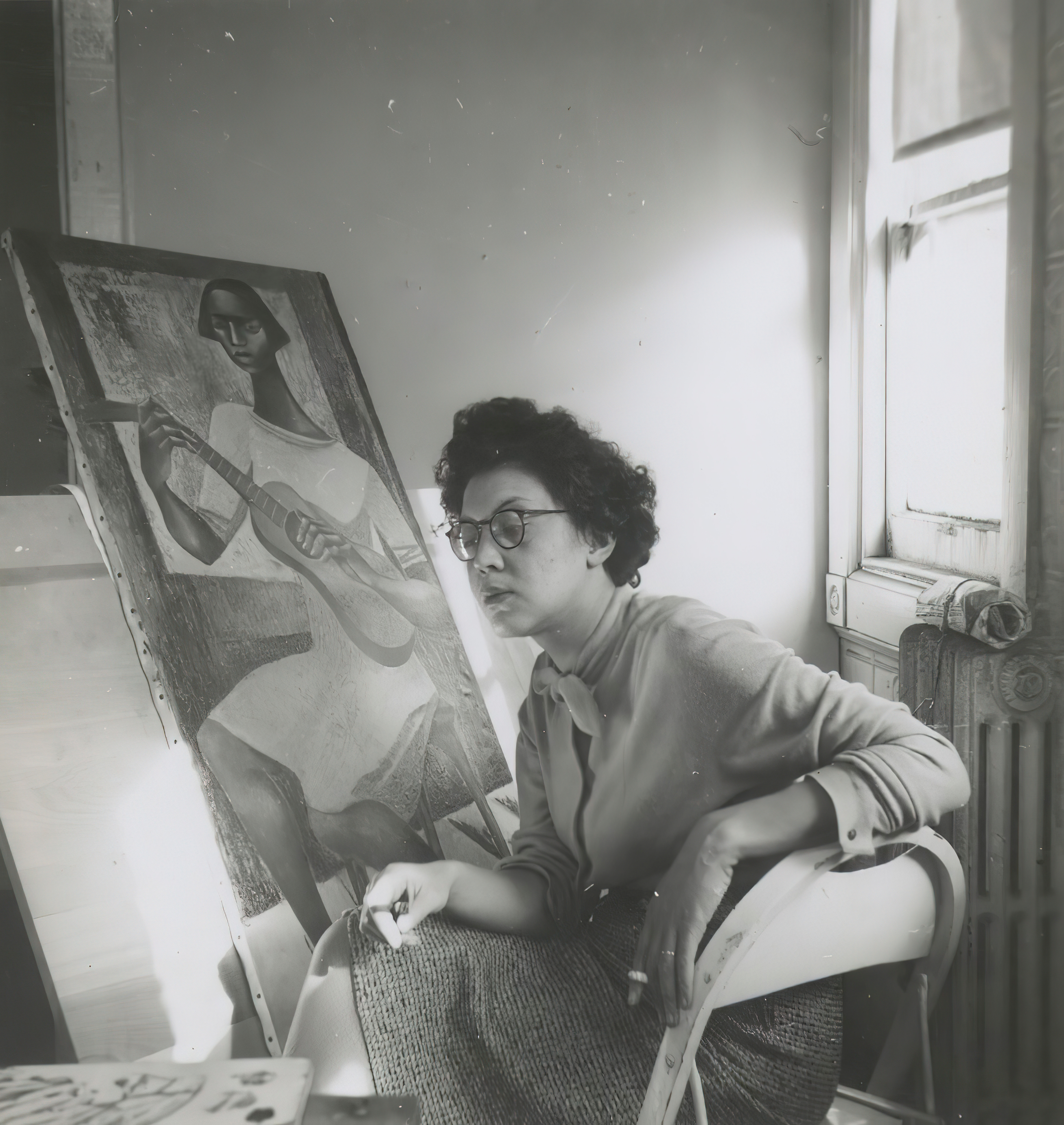 A black-and-white photo shows a young Elizabeth Catlett, medium-skinned with short, curly black hair, seated at an easel with her painting “Untitled (Woman in Yellow Hat).”