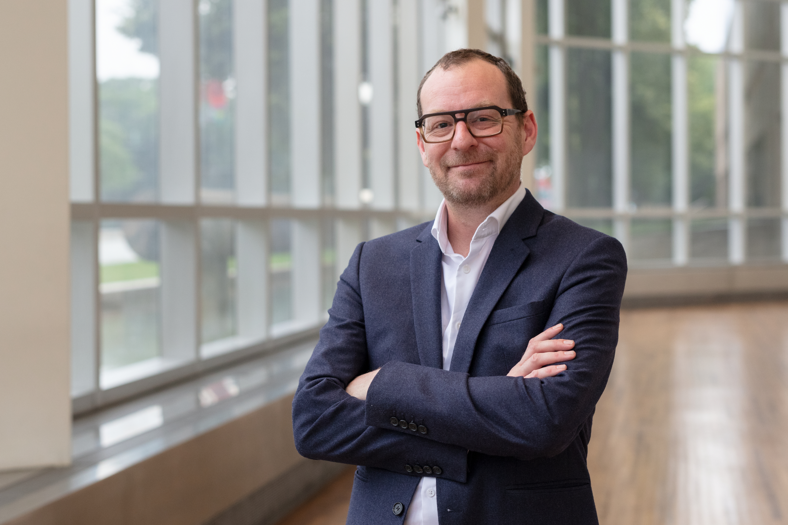 A light-skinned man in dark glasses and a suit jacket, Emerson Bowyer, stands with crossed arms and a slight smile in front of a long bank of glass windows.