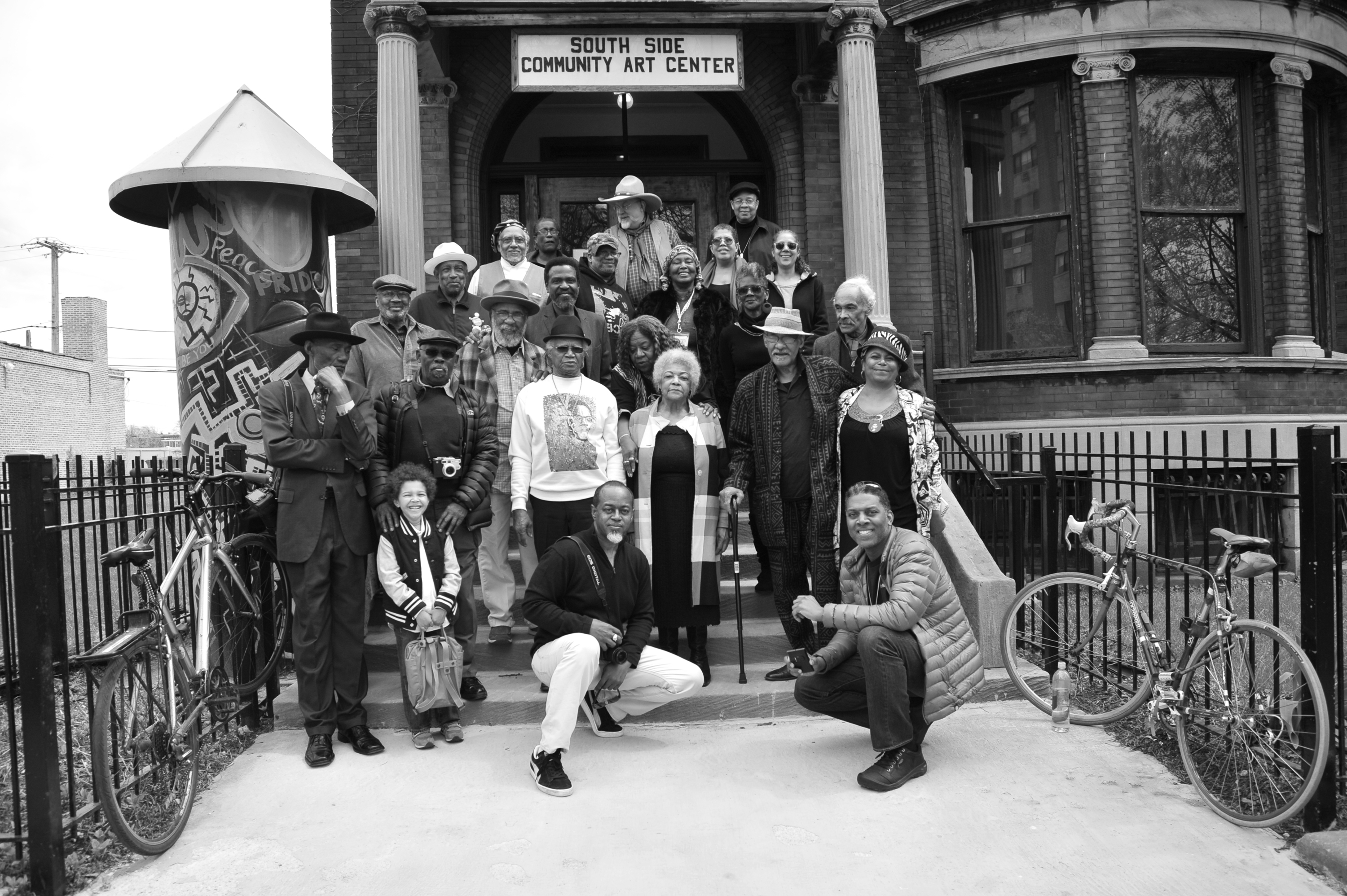 Group portrait in black and white showing over two dozen people of various ages and genders standing and sitting on the steps outside a historic brick building with a sign reading “South Side Community Art Center.” The group includes elders, middle-aged adults, and a few children, many wearing hats, sunglasses, or patterned garments. Two men kneel at the front holding cameras. On either side of the stairs, bicycles are parked against a metal fence. To the left, a cylindrical sculpture covered in colorful graffiti-style artwork stands beside the steps. The mood is celebratory and communal.