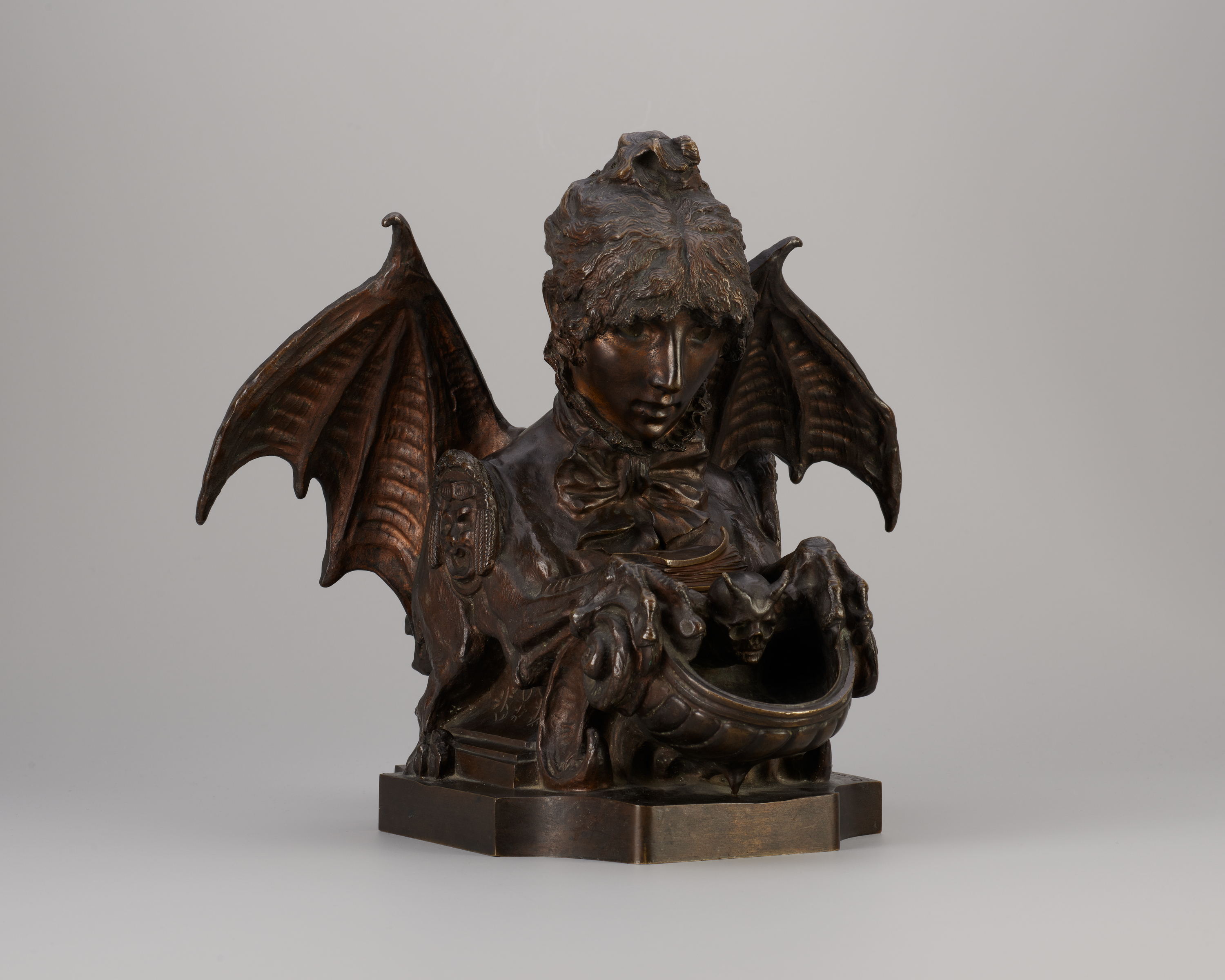 View in partial profile of a bronze-colored metal sculpture of a creature with the head of a curly-haired young woman, extended bat’s wings, and taloned claws. The figure looks down at a book in its lap.