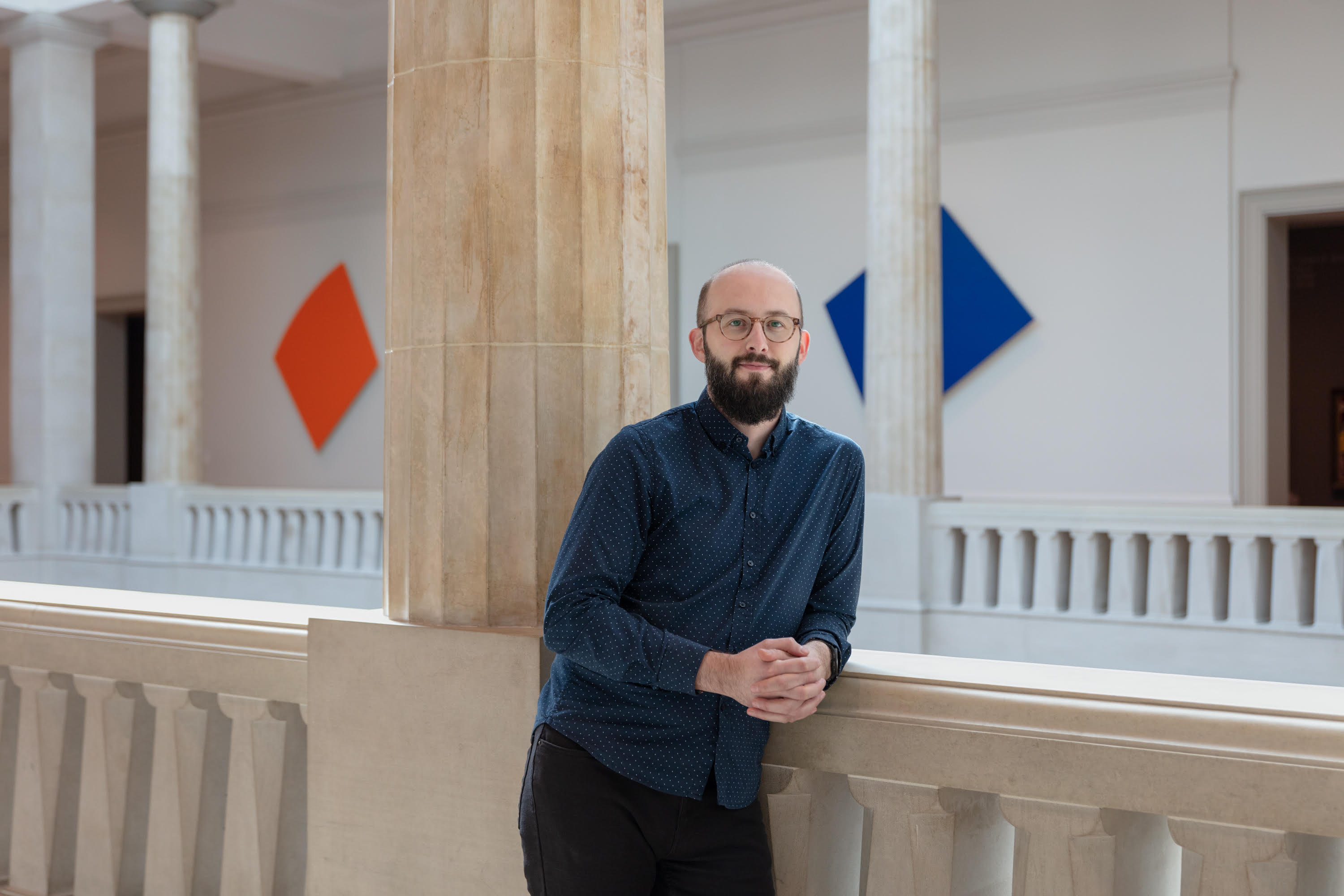 A light-skinned man with a full beard, a mustache, and round glasses —Kirill Mazor—smiles slightly as he leans upon a stone railing, quadrilateral artworks in single shades hung behind him.