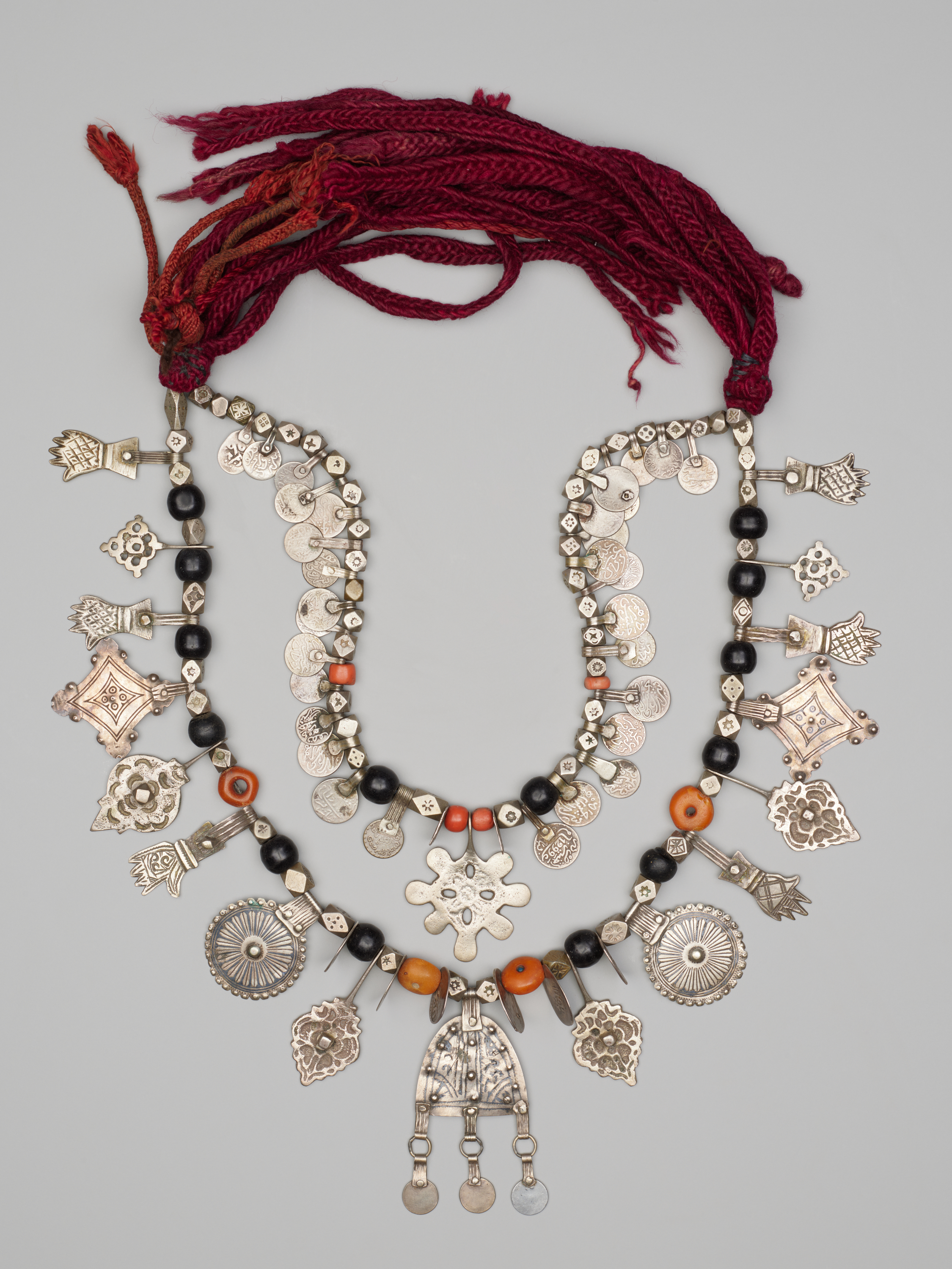A layered necklace on a light gray background: at top, red braided cords; below, two silver-beaded strands with black and orange beads, small coin discs, and many silver charms, including hand shapes at left and right and a bell-shaped pendant at bottom center.