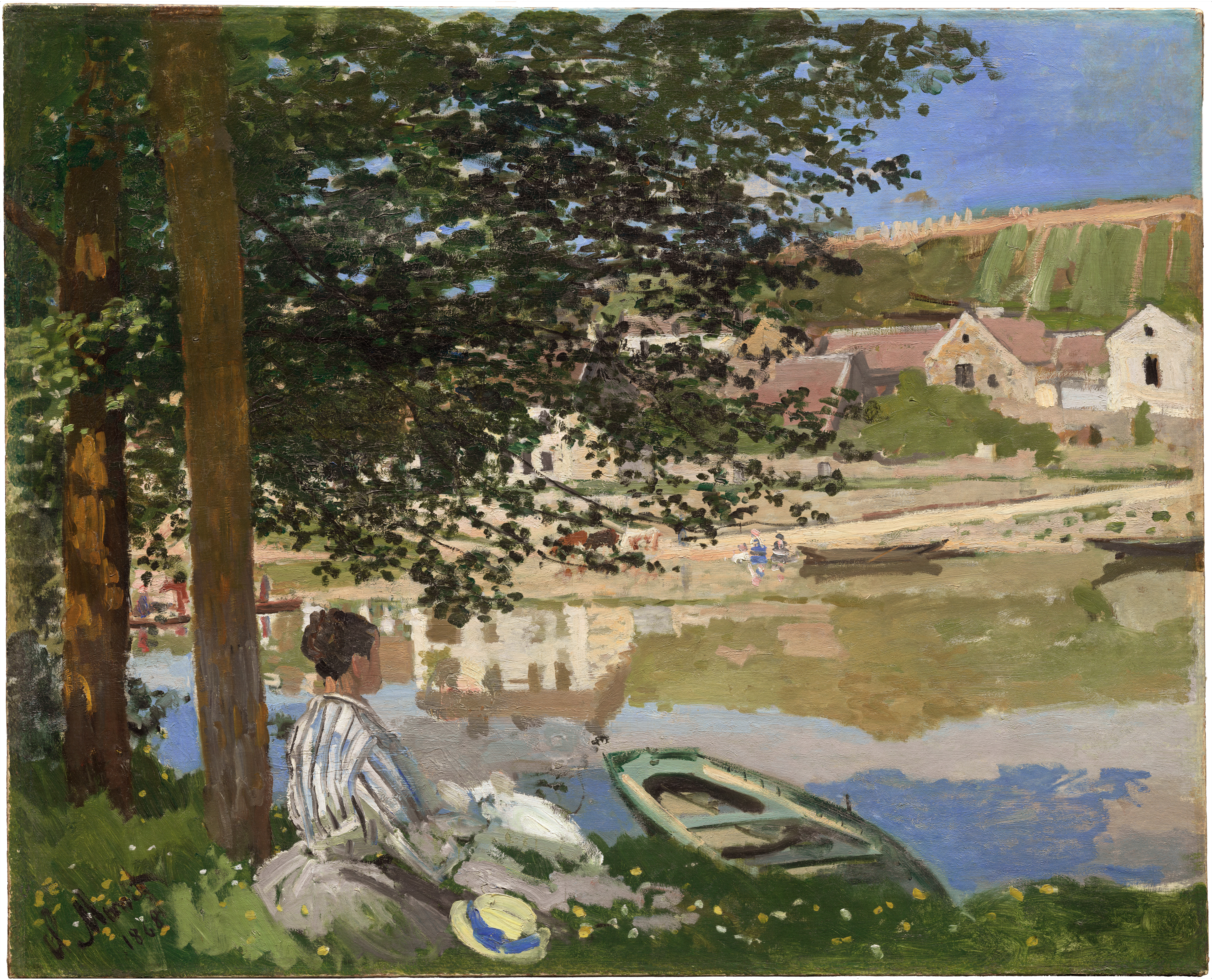 Foreground to background: On the left, tall brown tree trunks and dark green leaves shade a grassy bank where an adult with light skin and dark hair sits facing right, wearing a blue-and-white striped outfit; a round hat with yellow and blue bands lies nearby, and a green rowboat rests at the water’s edge. The center shows a calm river with reflections of trees and buildings, with two small figures on the opposite shore and a few narrow boats along the bank. In the distance, cream and tan houses with red roofs line the shore below a green hillside, topped by a clear blue sky.