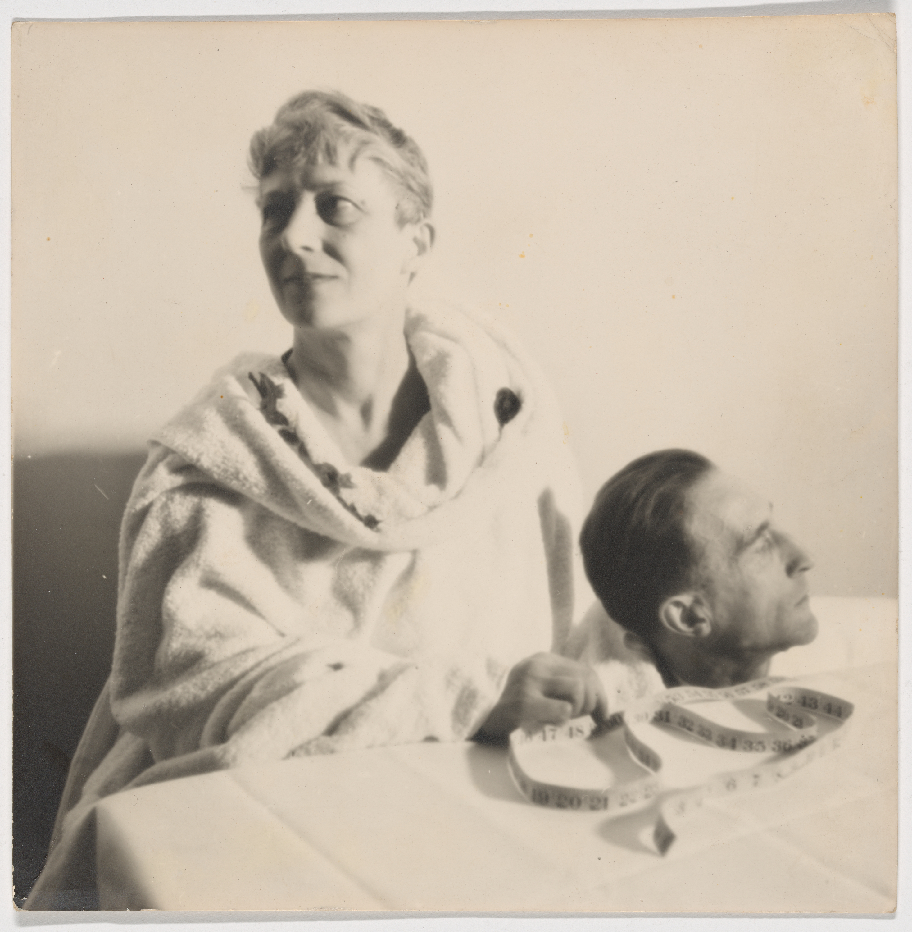 A sepia photograph of Mary Reynolds and Marcel Duchamp. Wearing a loose toga-like tunic, she sits at a table covered in a white cloth and looks up and to the right. Only Duchamp's head, looking off to his left, can be seen above the table. A flexible tape measure wraps around his neck; Reynolds holds the end of the tape measure.