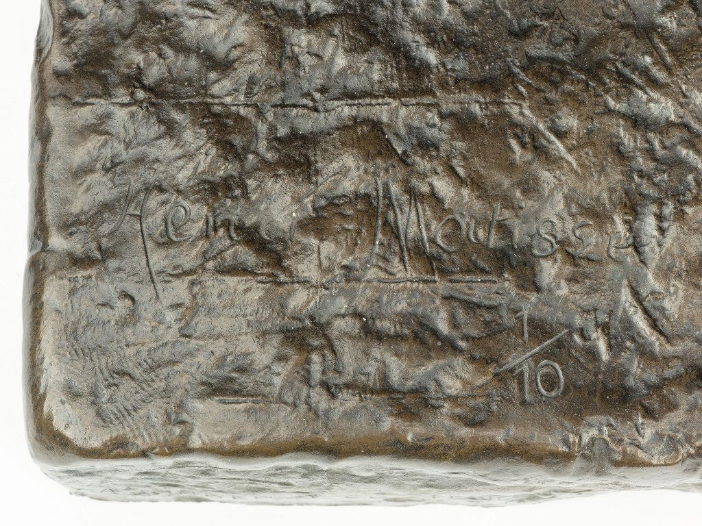 Top to bottom, a rough, dark brown surface with gray highlights fills the frame. Across the center, shallow incised text reads “Henri Matisse” from left to right. At the lower right, a small fraction “1/10” is etched. The bottom edge shows a slightly rounded corner and uneven texture.