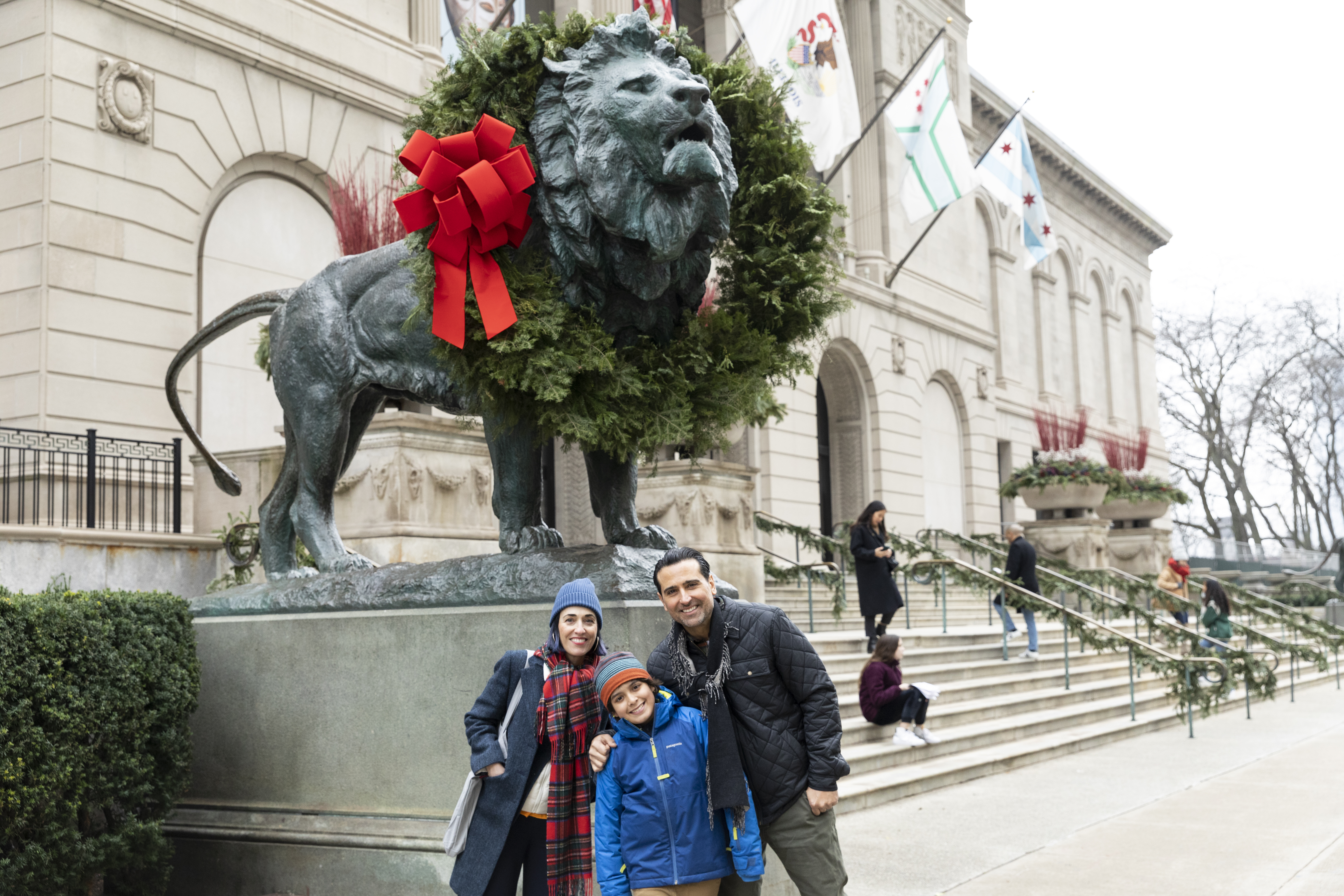 In a photo taken outside the Art Institute of Chicago, a family smiles in front of a large bronze lion that wears an evergreen wreath.