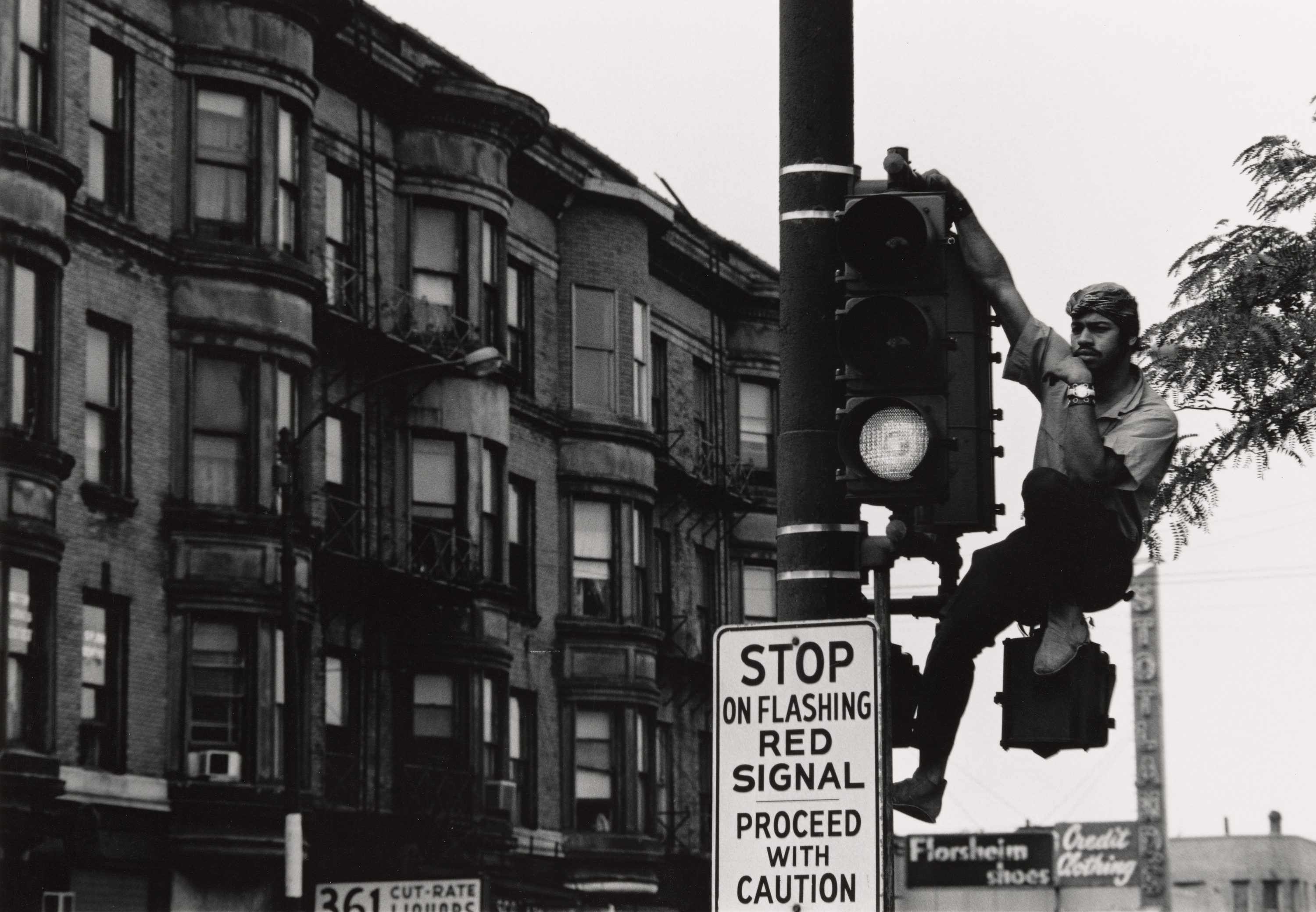 Black-and-white photograph of a man climbing and sitting on a traffic light pole in an urban street scene. He wears a headwrap, short-sleeved shirt, and pants, and looks thoughtfully into the distance with one hand resting on his chin. Below him is a sign that reads, “STOP ON FLASHING RED SIGNAL. PROCEED WITH CAUTION.” In the background are multi-story brick apartment buildings with bay windows and fire escapes, as well as visible storefront signs reading “361 CUT-RATE LIQUORS,” “Florsheim shoes,” and “Credit Clothing.”