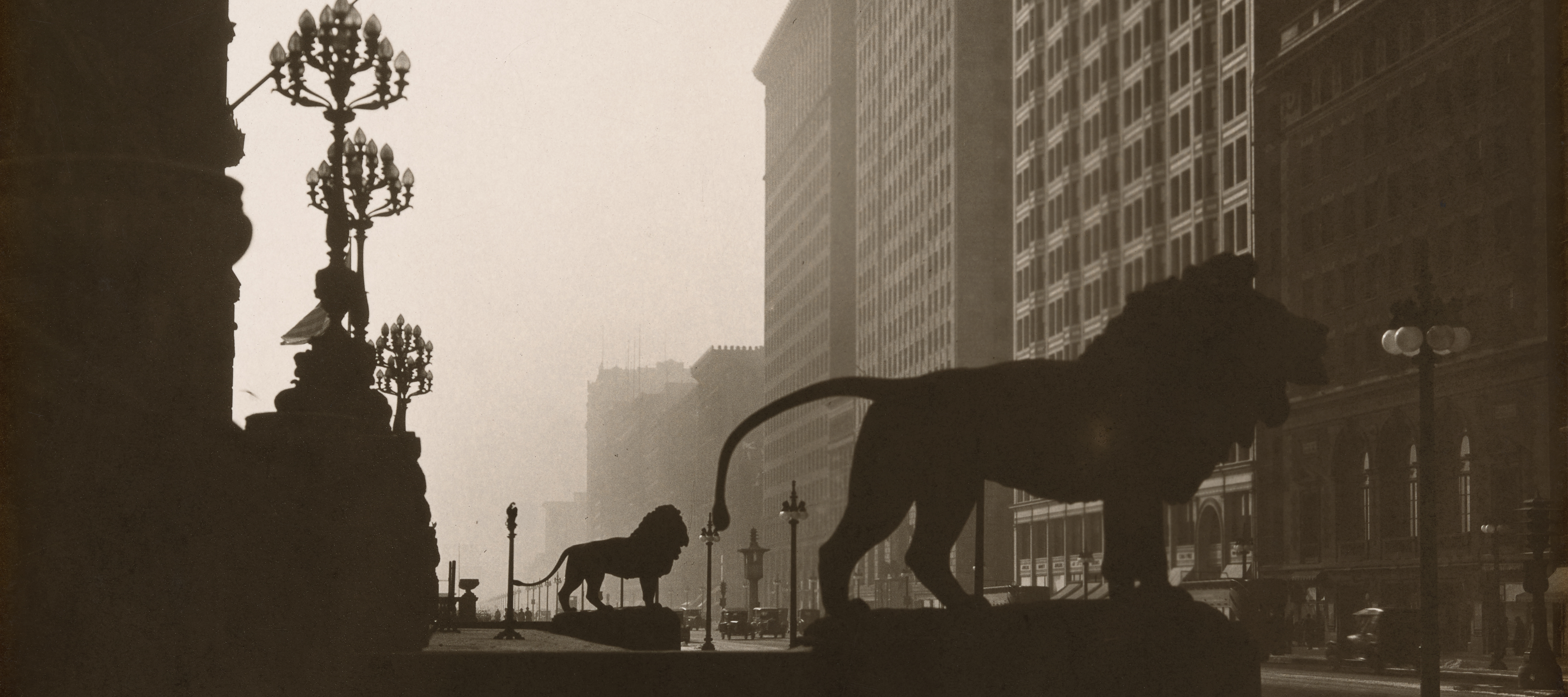 A black-and-white photo shows Michigan Avenue as viewed from just north of the steps of the Art Institute of Chicago. In the foreground, a massive stone lion looms in black silhouette. Beyond it is a second lion, also in silhouette. Behind them, to the left, are large lamps with tops that almost resemble chandeliers. In front of the lions and across the street, to the right, tall stone buildings rise to the top of the frame and beyond.
