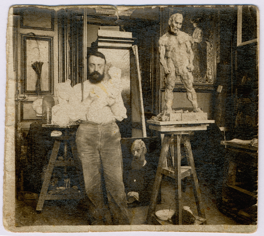 Top and back wall hold several framed pictures and a large rectangular panel. On the left, a person wearing a light shirt and dark pants stands beside pale sculptures on a worktable, and near the lower center another person sits close to the floor. On the right, a human figure sculpture in light tones stands on a tall wooden stand, about the same height as the standing person including the base, with small objects and a bowl on the floor nearby; overall colors are brown, beige, black, and white.