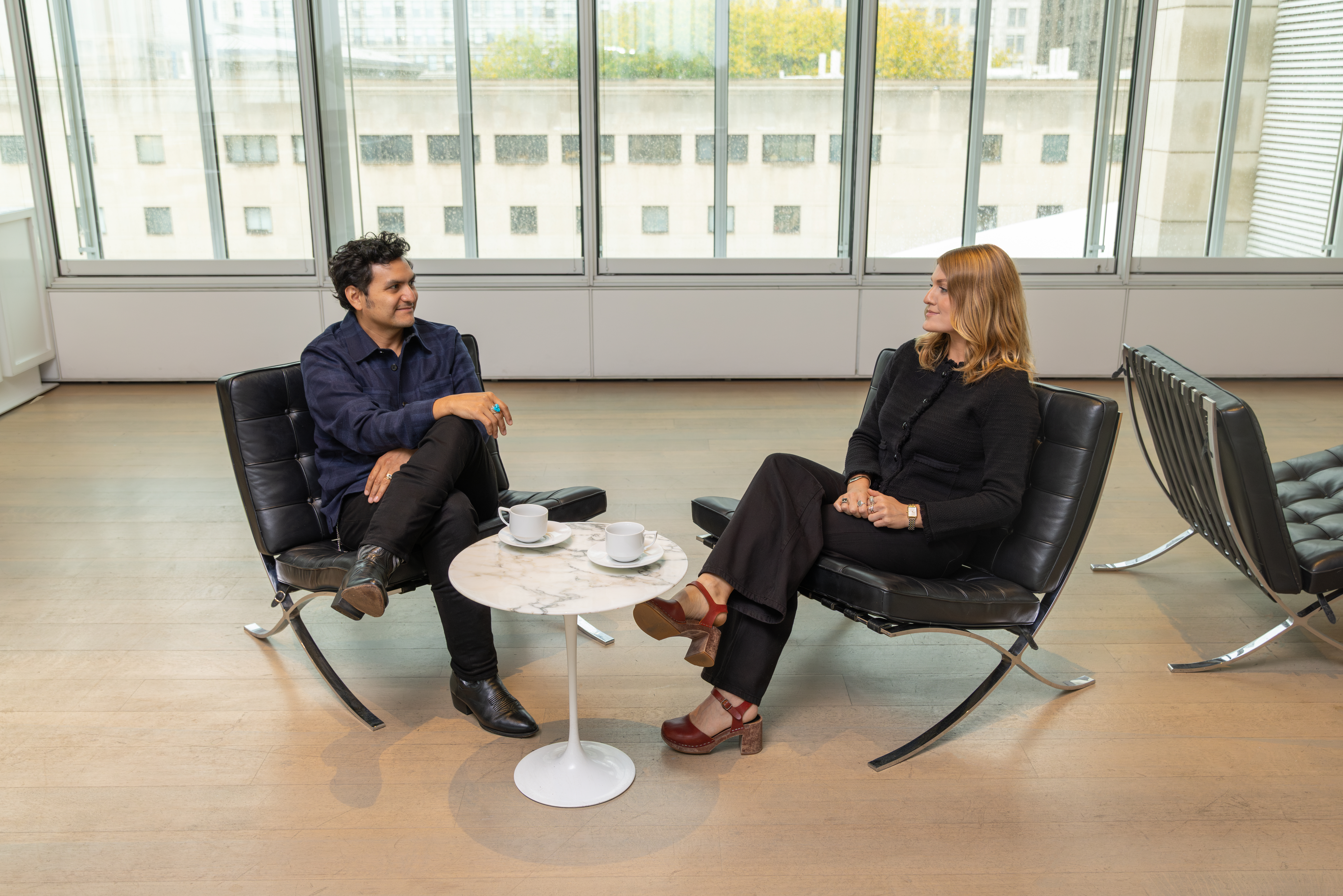 Sam Ramos, a medium-skinned man with dark hair wearing a dark-blue shirt, sits at left facing Maddie Shearer, a light-skinned blond woman in black, also seated. Between them is a small white table with cups of coffee.