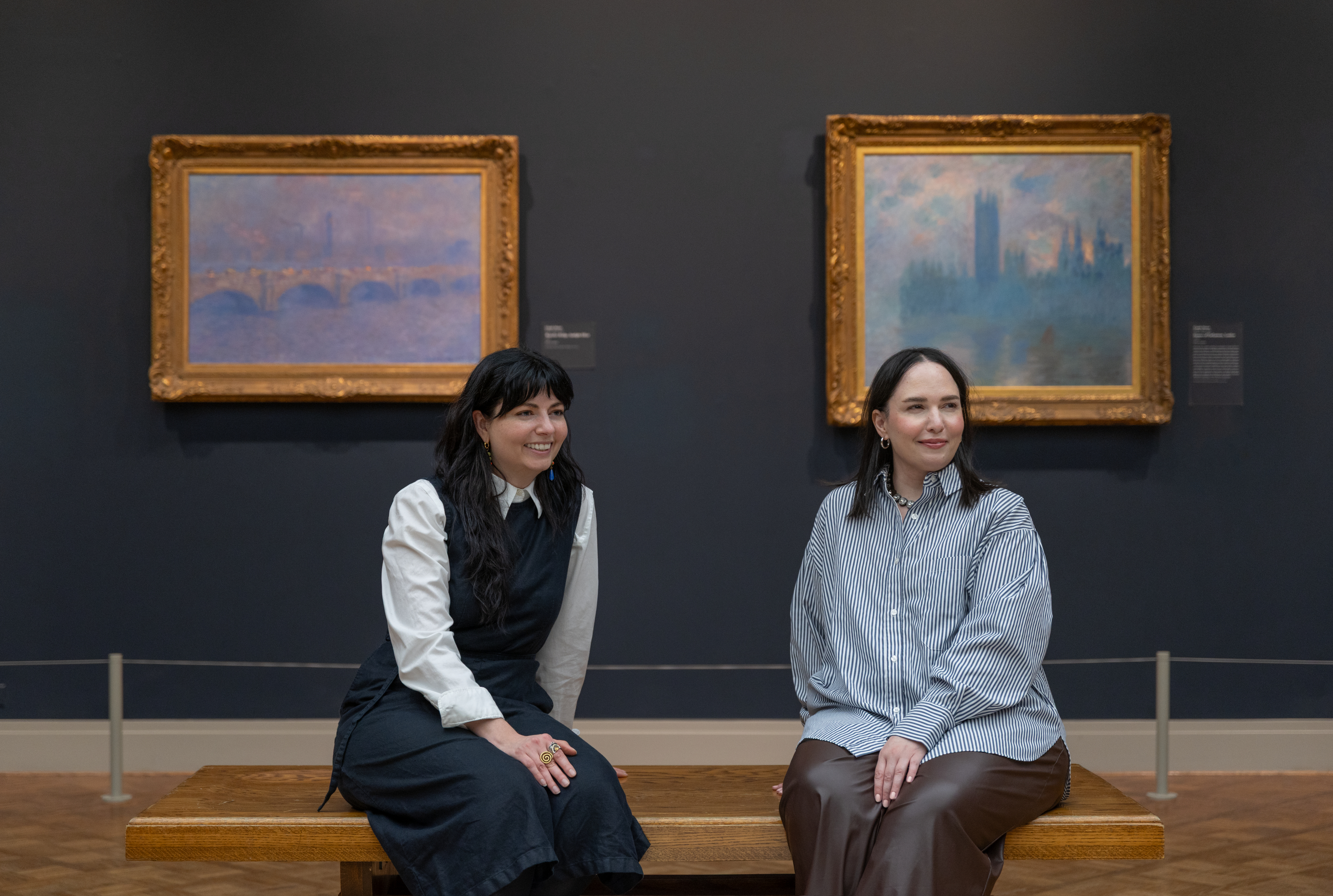 Color photograph of a museum gallery: at center, two women, Sam Grassi and Zahra Bahia, sit on a wooden bench; Sam wears a black dress over a white shirt and has long dark hair, and Zahra wears a gray striped blouse and brown skirt; at left and right, gold-framed blue paintings hang on a dark wall.