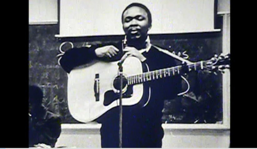 Black-and-white photograph of a man standing in front of a classroom chalkboard, holding an acoustic guitar. He is positioned behind a microphone and appears to be performing or speaking. He wears a dark sweater over a collared shirt and has a focused but calm expression. The guitar strap crosses his chest as his left hand holds the guitar neck and his right hand rests near the tuning pegs. A second person is partially visible seated in the lower left corner. The background includes a chalkboard with faint writing and a projection screen above it.