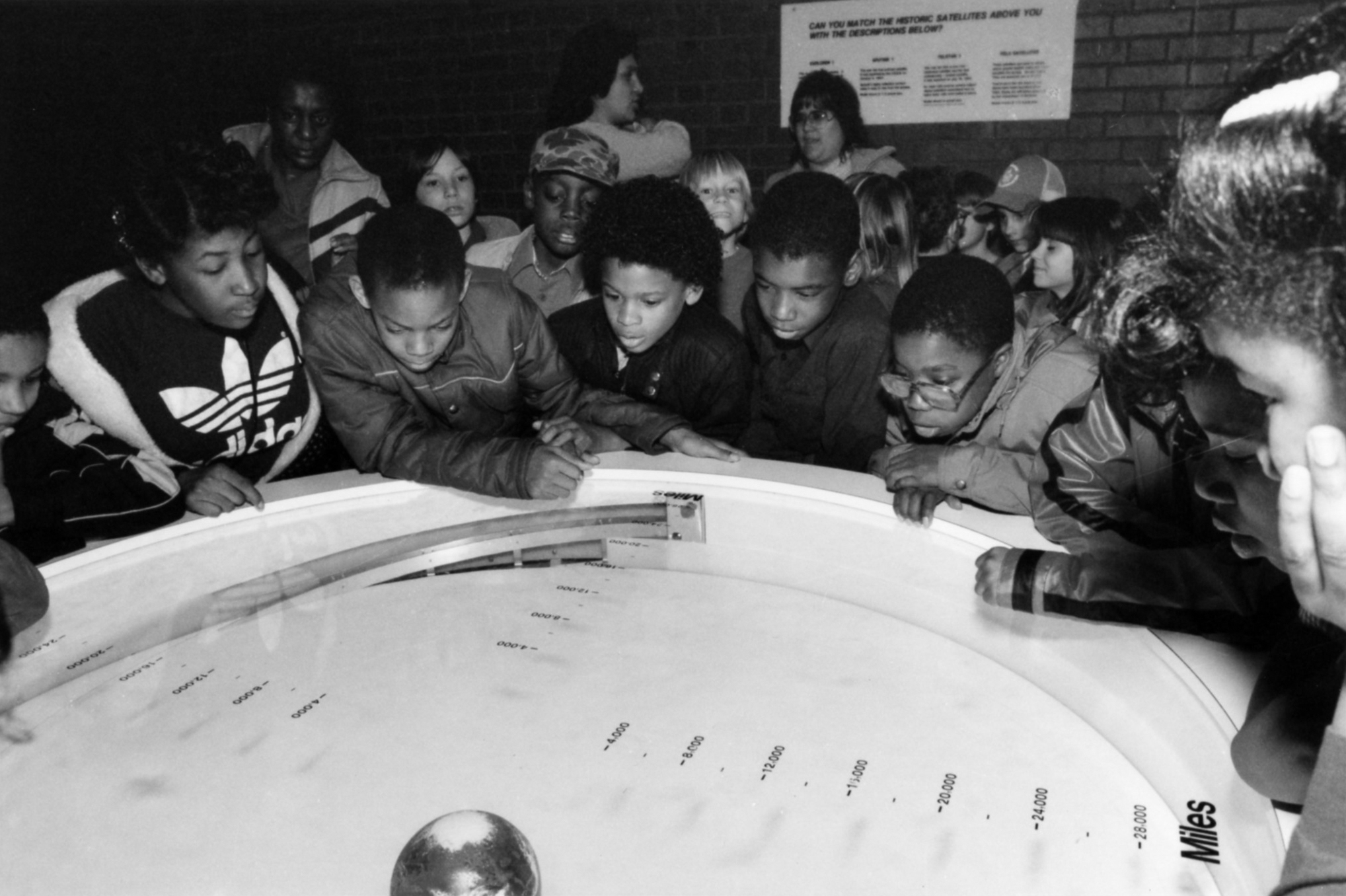 A grainy black-and-white photograph of a group of children crowding around a circular display. They lean on and over it, observing and reading.